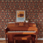 An artistic setting featuring an old wooden piano against a vibrant background of Morris Autumn Wallpaper. The wallpaper has a detailed pattern of red and beige floral motifs with navy accents, giving a classic and rich ambiance to the space.