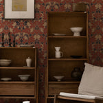 A cozy corner of a room showcasing a wooden bookshelf filled with ceramics and home decor items. The backdrop is the Morris Autumn Wallpaper, with its intricate floral patterns in shades of red, navy, and beige, complementing the earthy tones of the room's furnishings.