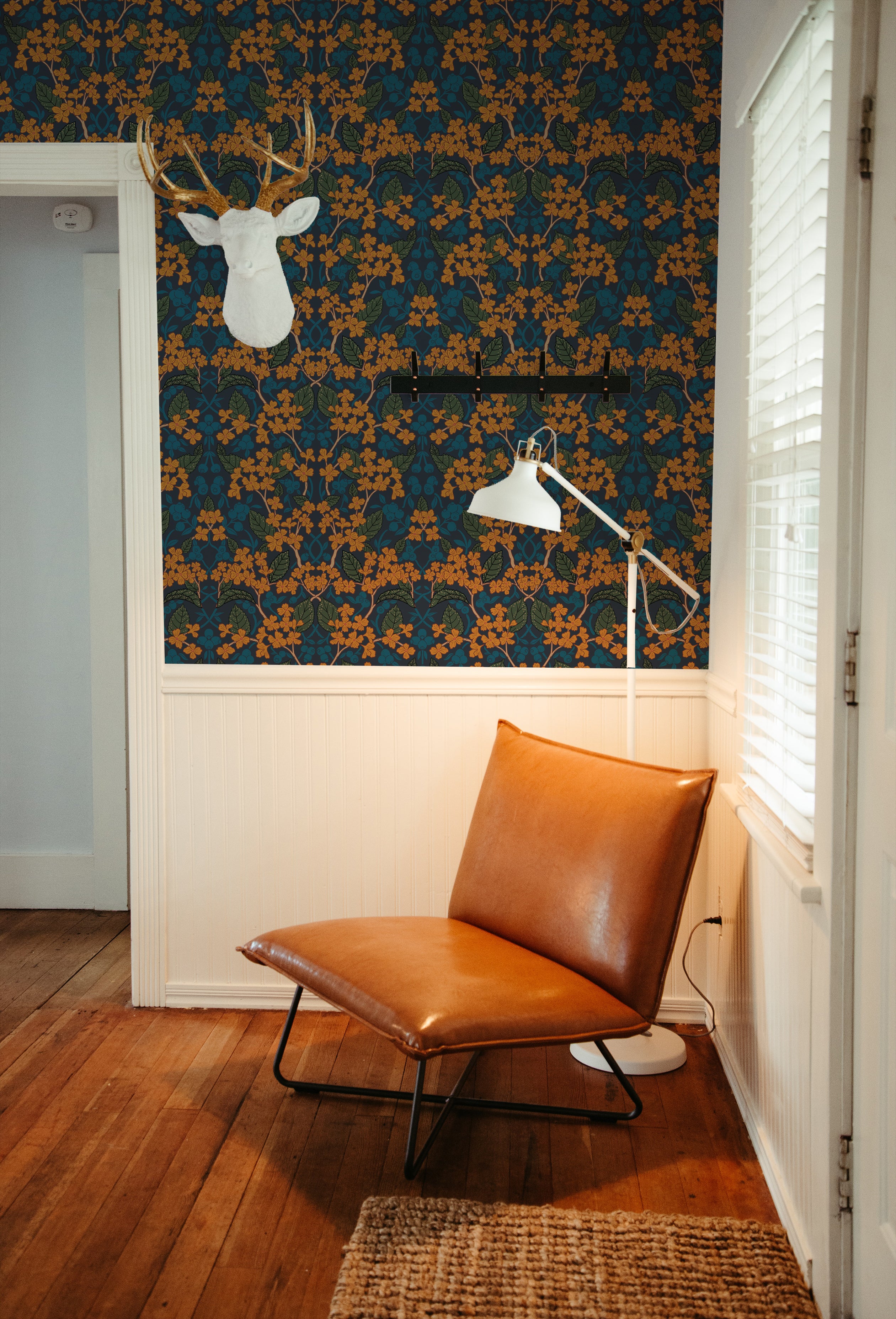 A cozy reading nook featuring Autumn Boho wallpaper with a floral pattern in shades of orange, blue, and green, accentuated by a white deer head sculpture, a modern leather sling chair, and a wooden floor