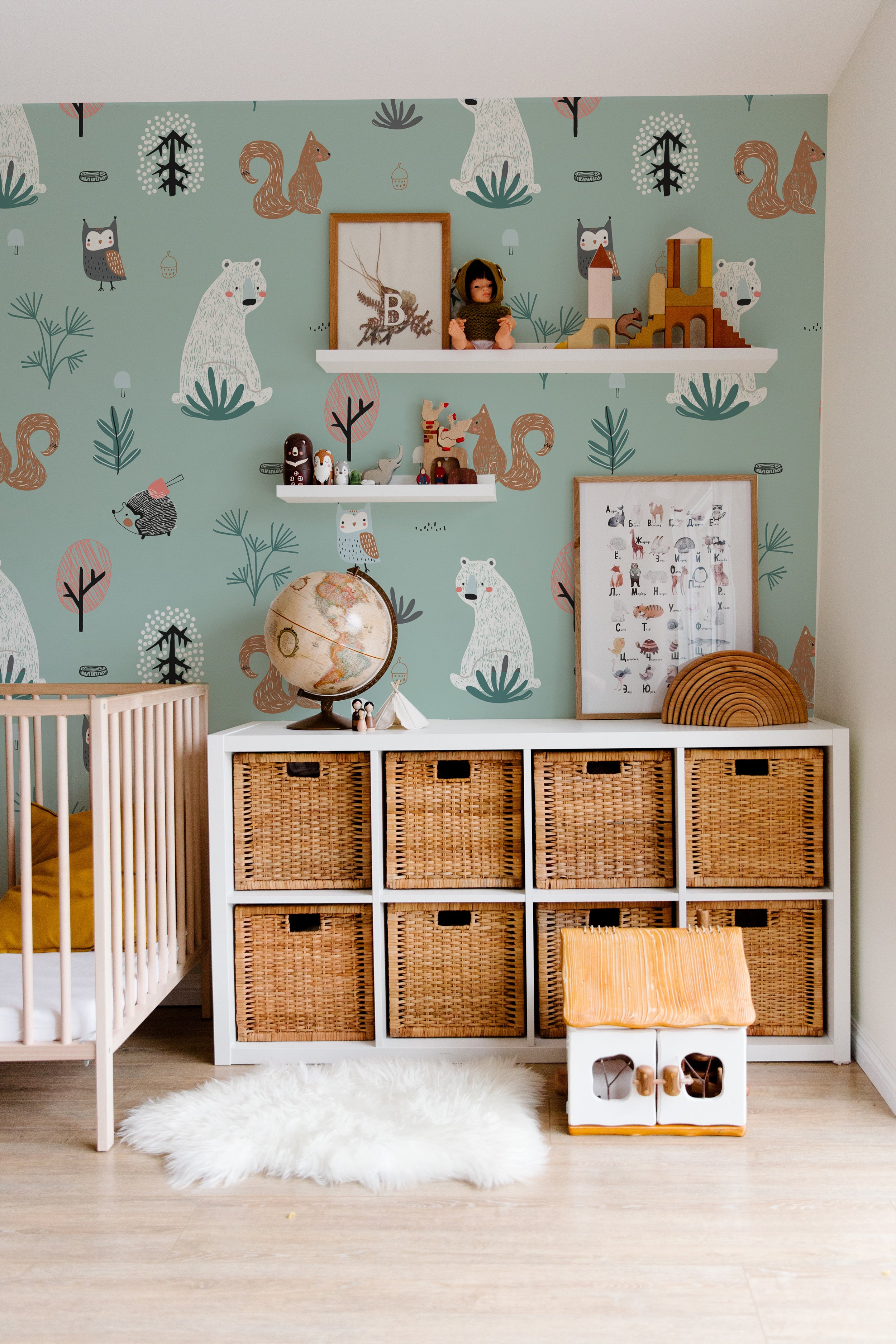 This nursery room is beautifully adorned with "Kids Wallpaper - Forest Critters - 50 inches." The wallpaper features large prints of friendly woodland creatures such as bears, owls, and squirrels in a whimsical forest setting. The room is furnished with a white crib, rattan storage baskets, and playful decor, creating a charming and inviting space for children.