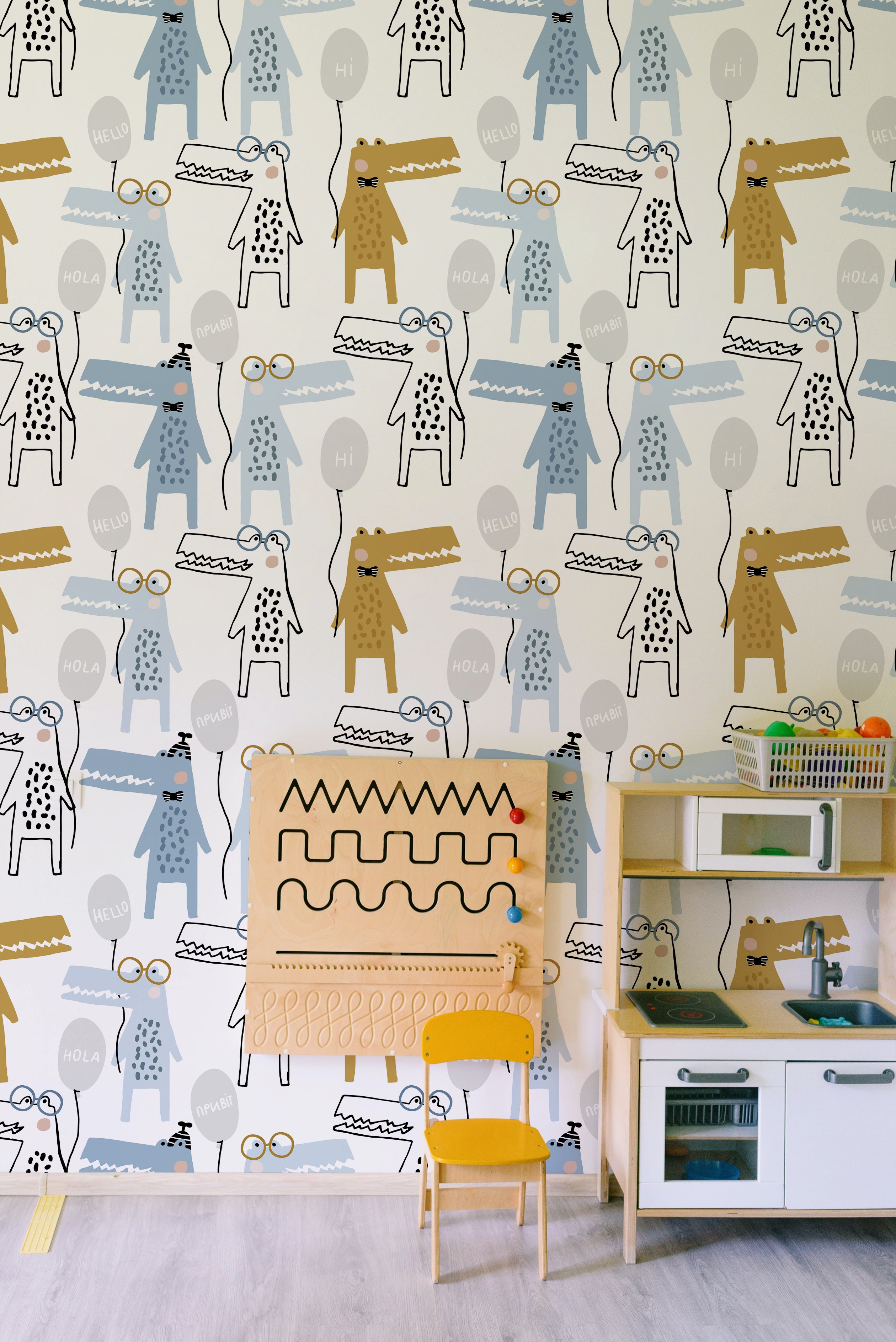 A playful children's room interior decorated with the "Party Crocs Wallpaper - 25 inches." The room includes a wooden play kitchen, a yellow chair, and a sensory board. The wallpaper adds a fun, creative touch with its colorful crocodile characters and friendly greetings in various languages.