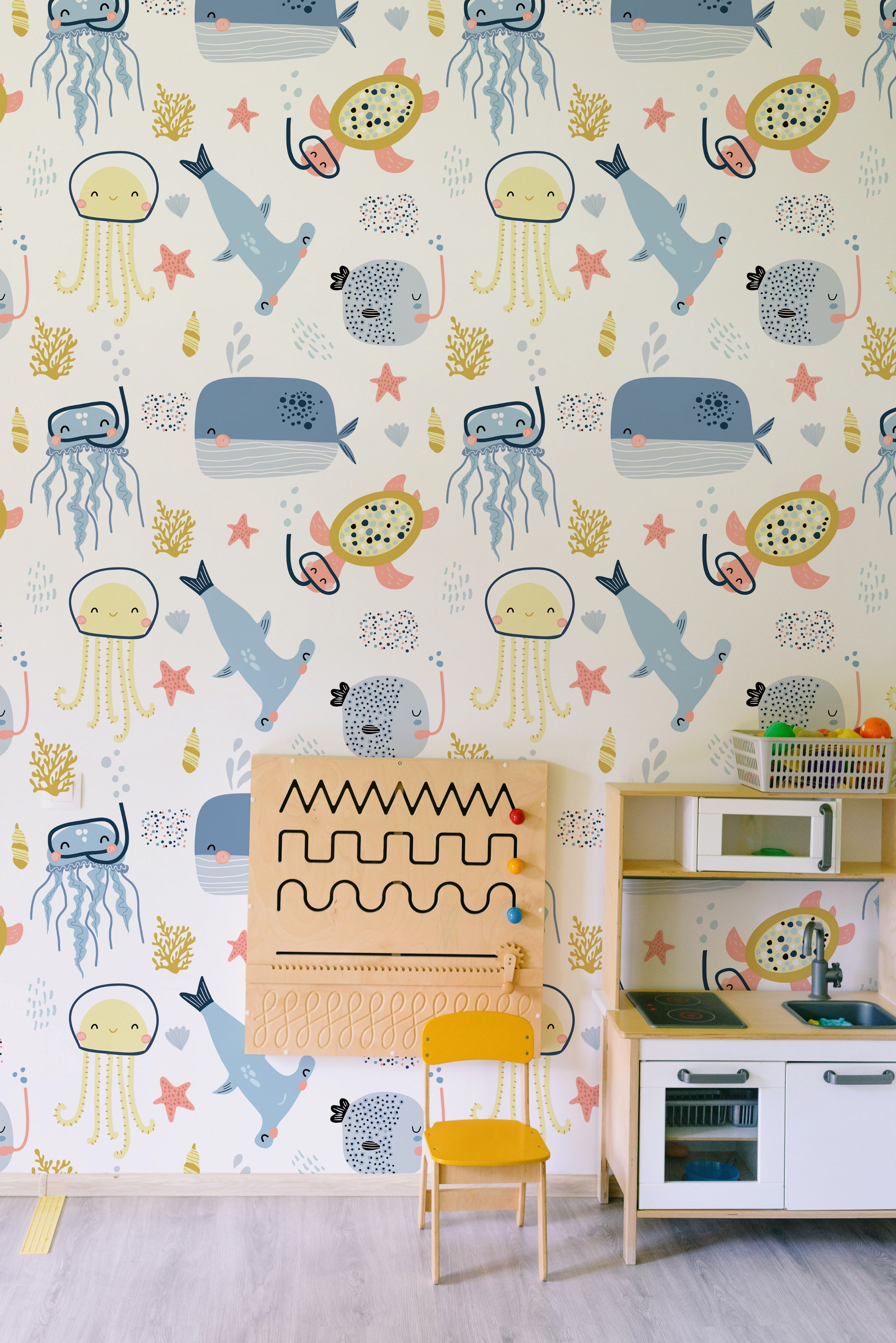  A colorful children's room featuring a wall adorned with an under-the-sea-themed wallpaper. The wallpaper includes playful illustrations of sea creatures like jellyfish, whales, sea turtles, and coral. The room is furnished with a small play kitchen and a wooden activity board, creating an engaging and imaginative space.