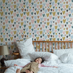 A child lying peacefully on a bed with white bedding in front of a wall decorated with the Amore Wallpaper. The wallpaper features a pattern of multicolored hearts in shades of blue, pink, yellow, and black, creating a playful and loving atmosphere suitable for a child's bedroom.