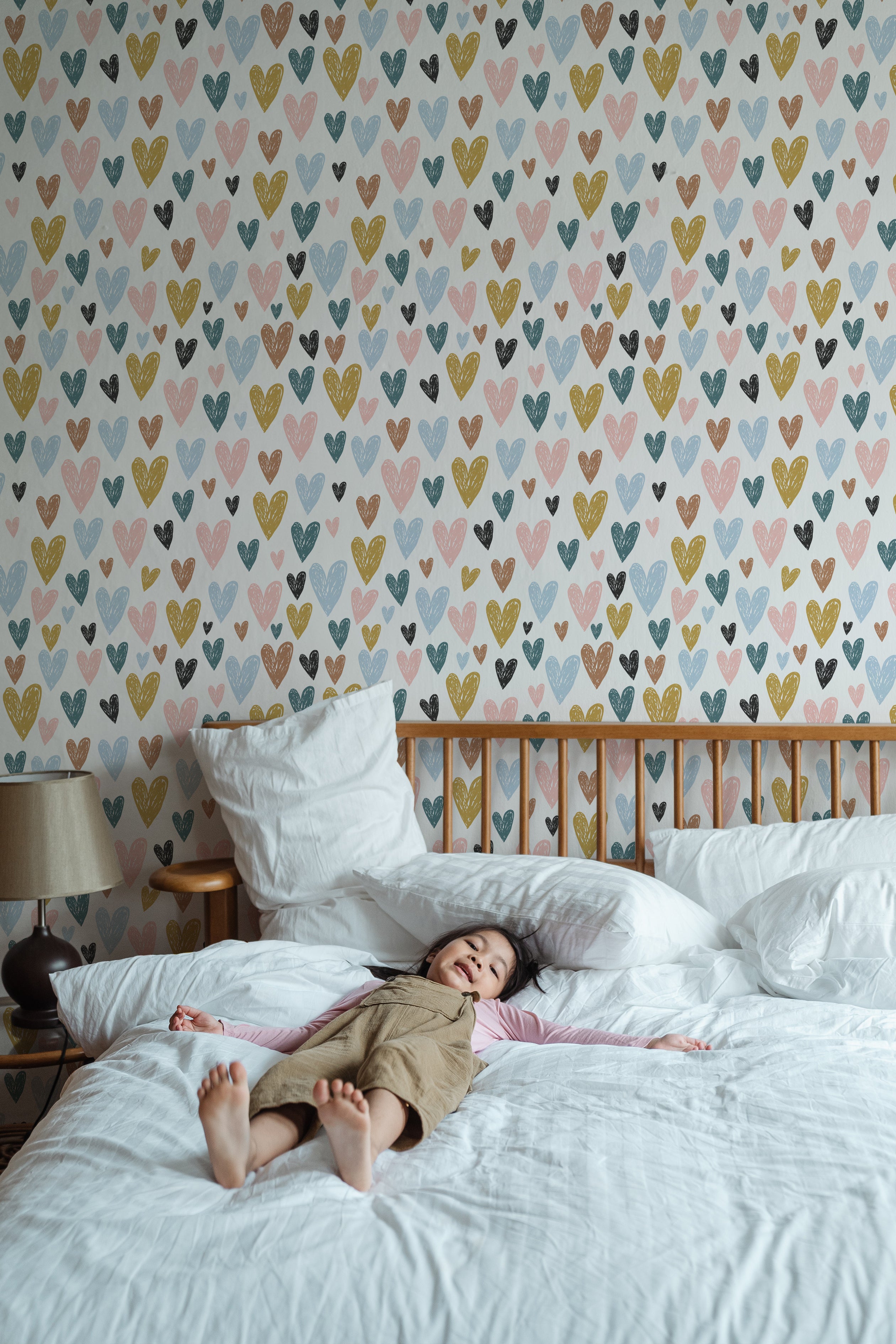 A child lying peacefully on a bed with white bedding in front of a wall decorated with the Amore Wallpaper. The wallpaper features a pattern of multicolored hearts in shades of blue, pink, yellow, and black, creating a playful and loving atmosphere suitable for a child's bedroom.