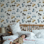 A young child sleeps peacefully on a large white bed in a room decorated with Toy Cars Wallpaper. The playful wallpaper features a variety of stylized cars in shades of blue, yellow, and gray, scattered playfully across a light background