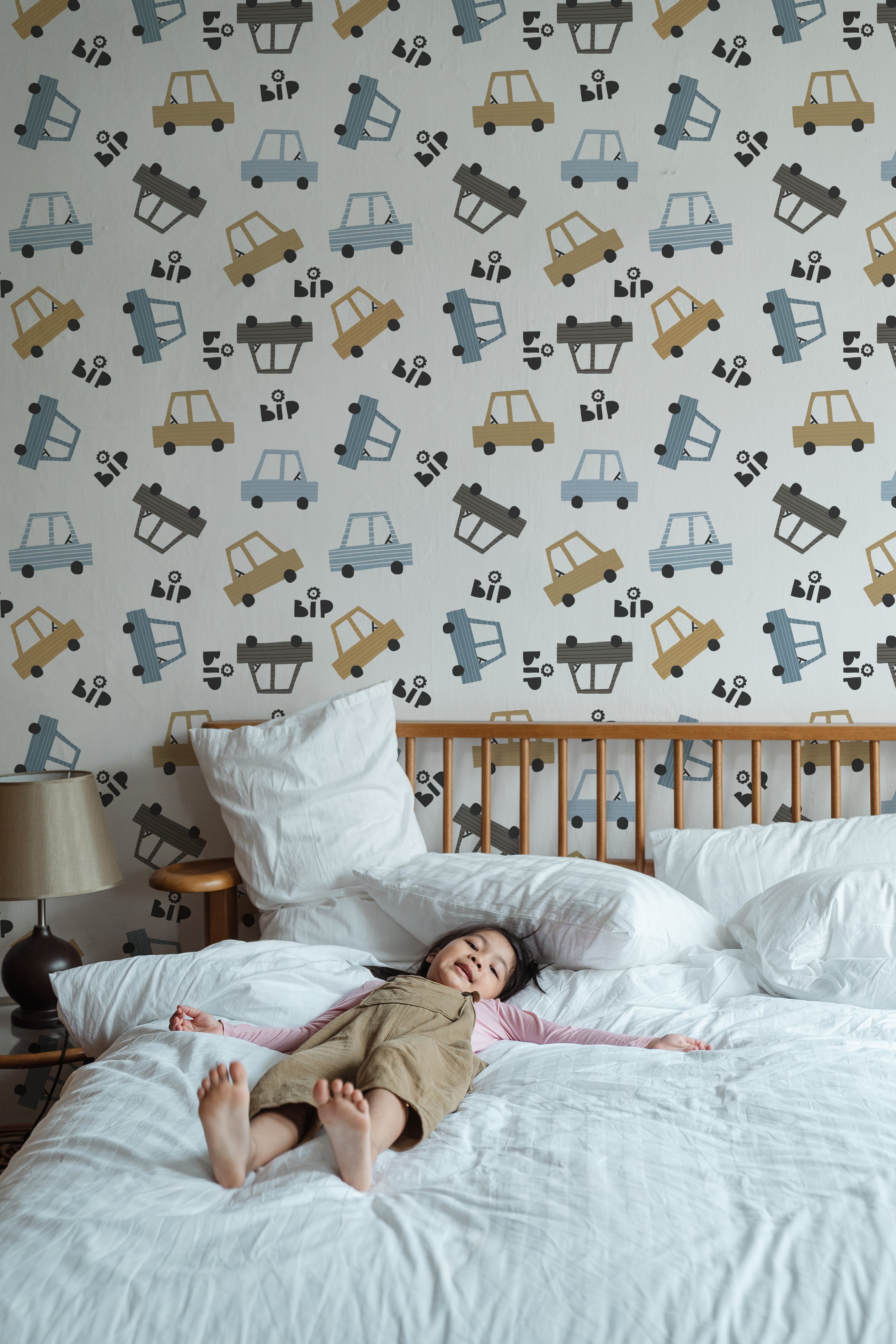 A young child sleeps peacefully on a large white bed in a room decorated with Toy Cars Wallpaper. The playful wallpaper features a variety of stylized cars in shades of blue, yellow, and gray, scattered playfully across a light background