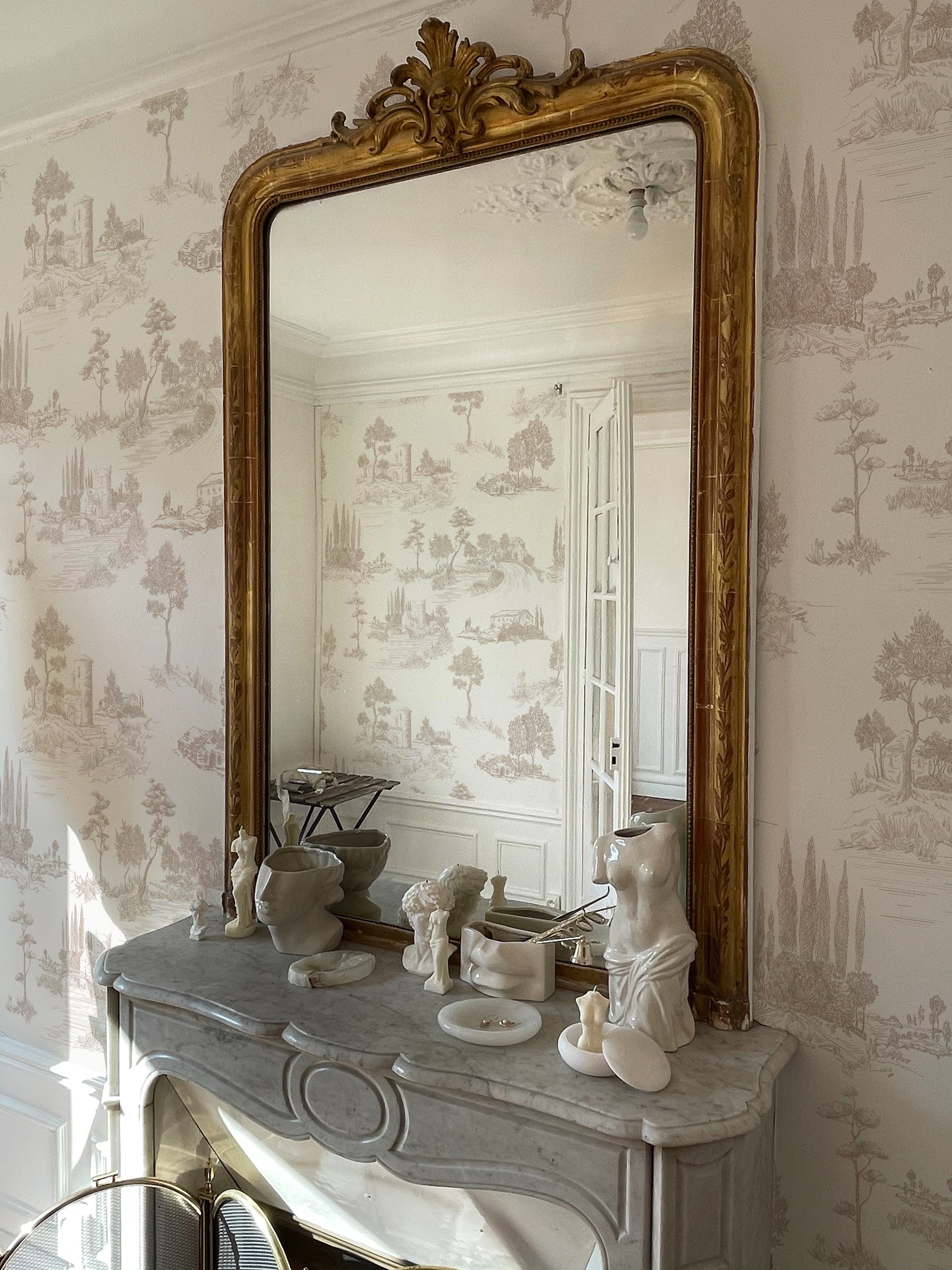 An elegant interior showcasing the Farmhouse Toile Wallpaper on the wall behind an ornate golden-framed mirror, sitting above a marble fireplace adorned with sculptural art pieces, reflecting the wallpaper's classic design and creating a sophisticated vintage aesthetic.