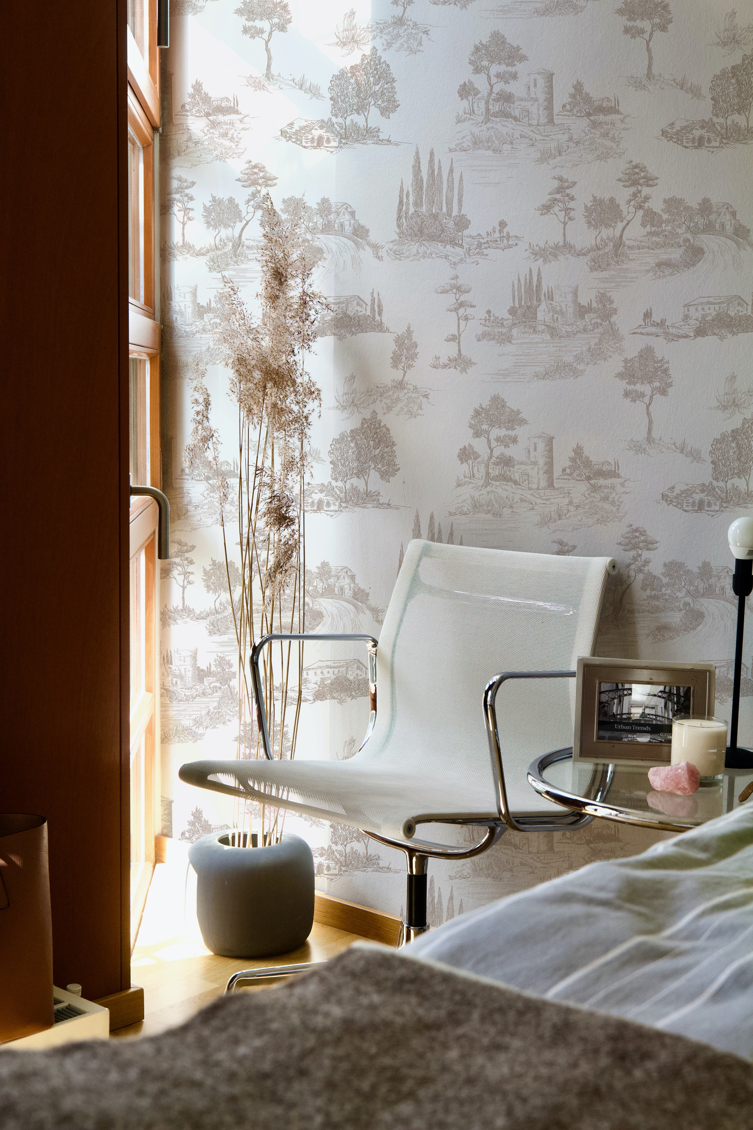A cozy bedroom corner illuminated by warm sunlight, with the Farmhouse Toile Wallpaper providing a quaint and picturesque backdrop to a modern chair and side table, blending contemporary furnishings with the timeless allure of the toile pattern.