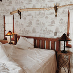 A rustic bedroom setting featuring the Farmhouse Toile Wallpaper, which complements the wooden bed frame and traditional decor, highlighting the wallpaper's ability to infuse historical elegance into a homely and inviting atmosphere.