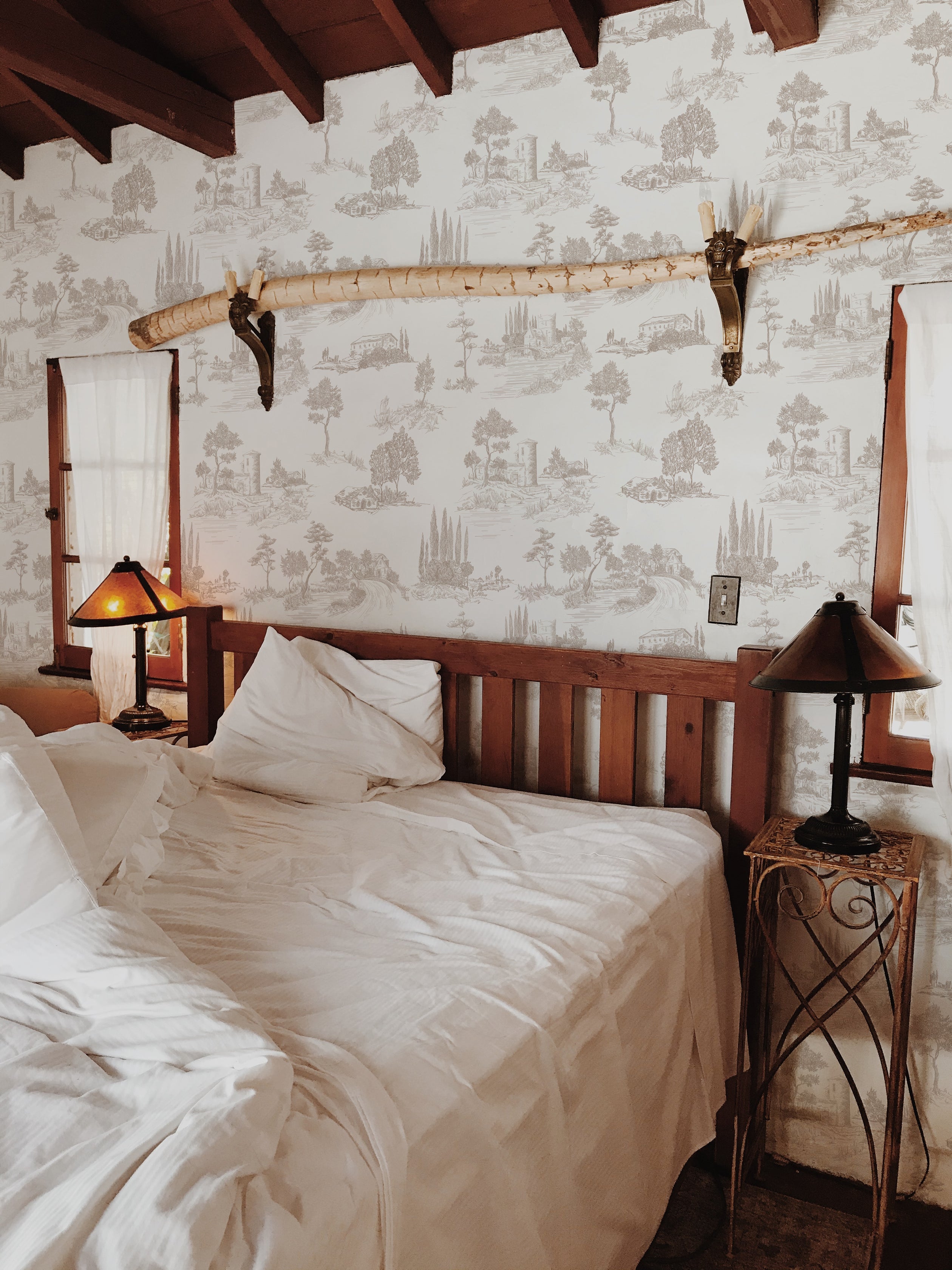 A rustic bedroom setting featuring the Farmhouse Toile Wallpaper, which complements the wooden bed frame and traditional decor, highlighting the wallpaper's ability to infuse historical elegance into a homely and inviting atmosphere.