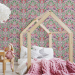 A cozy child's bedroom featuring a house-shaped bed frame made of natural wood, set against a vibrant floral and fruit-patterned wallpaper with a pink background. The room includes a thick, chunky pink blanket, white pillows, and a teddy bear seated on a wooden chair."
