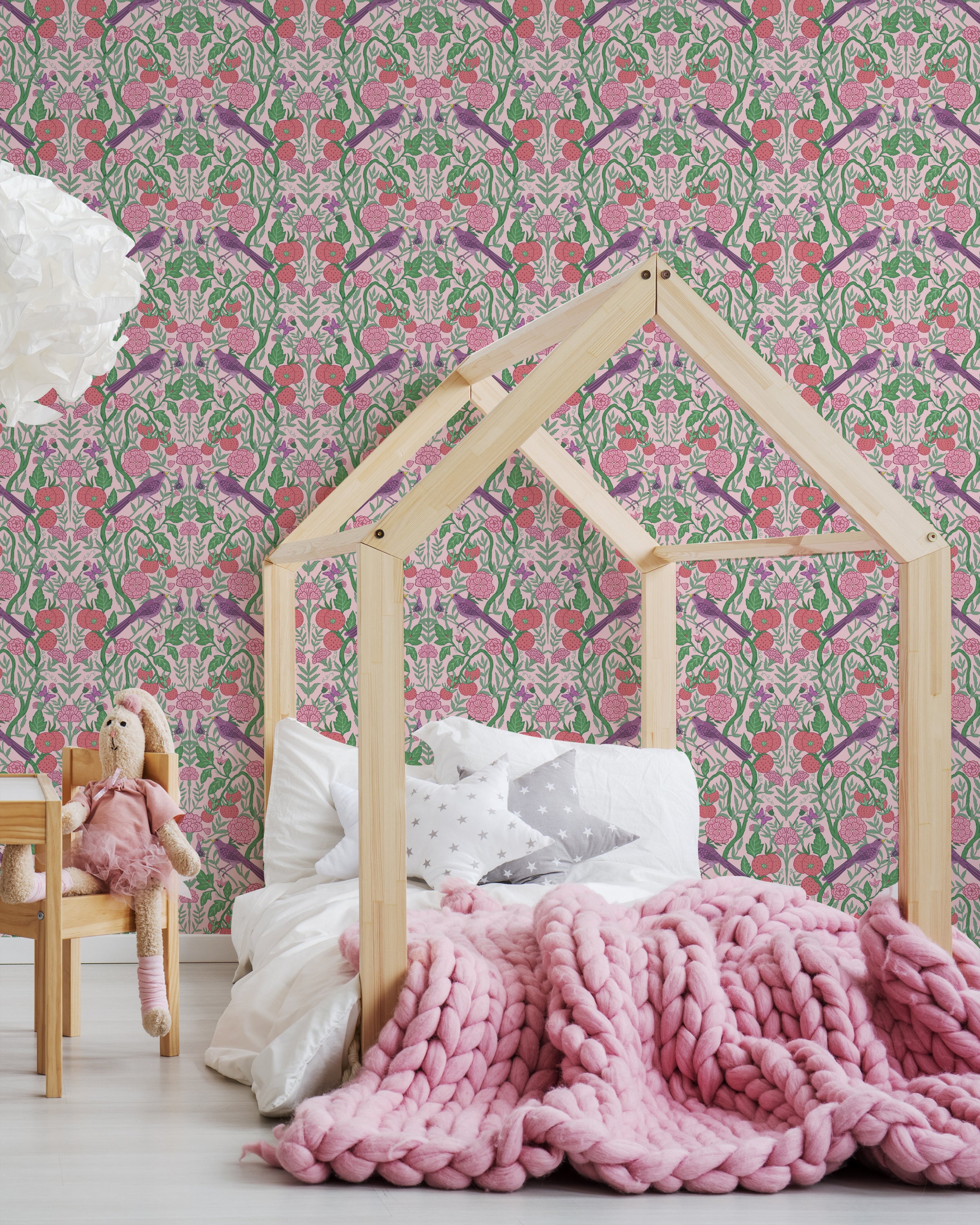 A cozy child's bedroom featuring a house-shaped bed frame made of natural wood, set against a vibrant floral and fruit-patterned wallpaper with a pink background. The room includes a thick, chunky pink blanket, white pillows, and a teddy bear seated on a wooden chair."