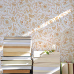 A stack of hardcover books in various sizes leans against a wall covered with De Pijp Floral Wallpaper, which features a golden floral pattern on a light background. The pattern includes stylized sunflowers and delicate fern-like plants, catching the light that streams through a nearby window.