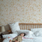 A young child lies relaxed on a large bed in a bedroom adorned with De Pijp Floral Wallpaper. The wallpaper showcases a soft golden floral design on a cream background, creating a serene and cheerful atmosphere. The room is furnished with a wooden bed and a vintage bedside lamp.
