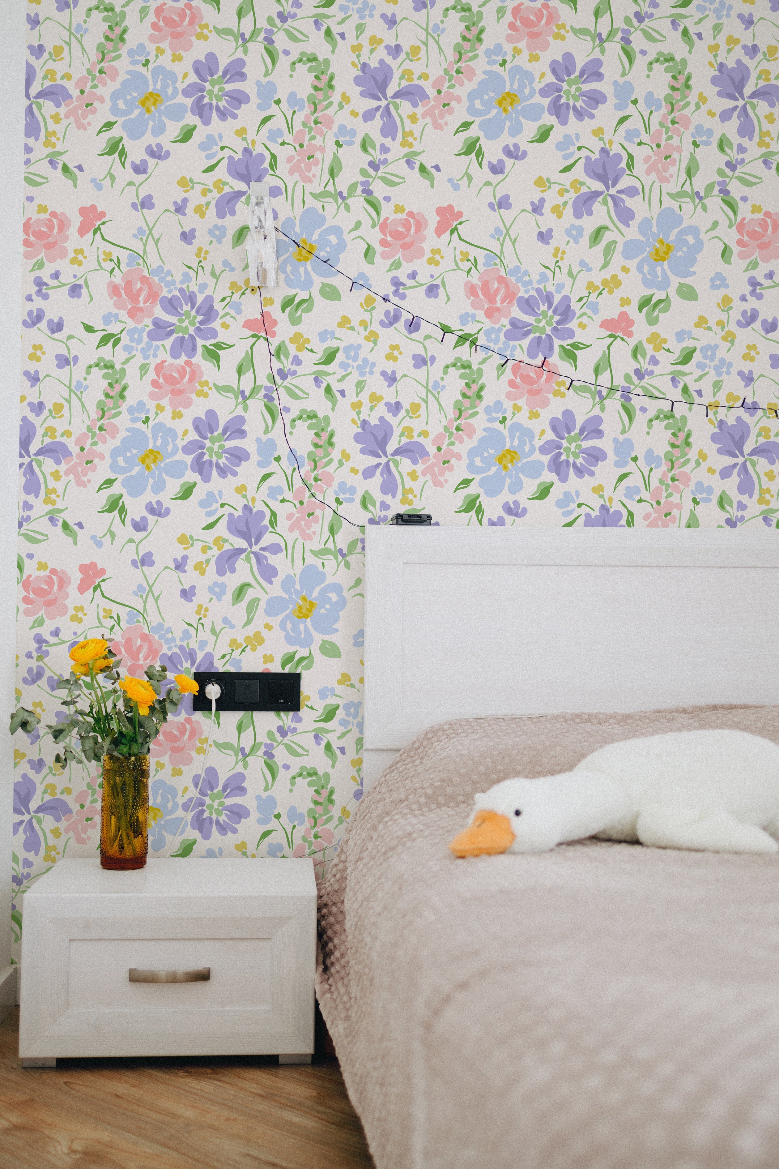 A cozy bedroom corner with Romana Wallpaper displaying a vibrant floral pattern of pink, yellow, and blue flowers on a light cream background. A small white nightstand with books and a vase of fresh yellow roses enhances the room's fresh, inviting feel.