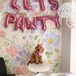 A festive birthday setting for a dog named 'Pawty' with colorful balloons spelling 'Let's Pawty Happy Birthday' against a backdrop of Romana Wallpaper. The wallpaper features a cheerful floral pattern in pastel shades of pink, blue, and yellow.