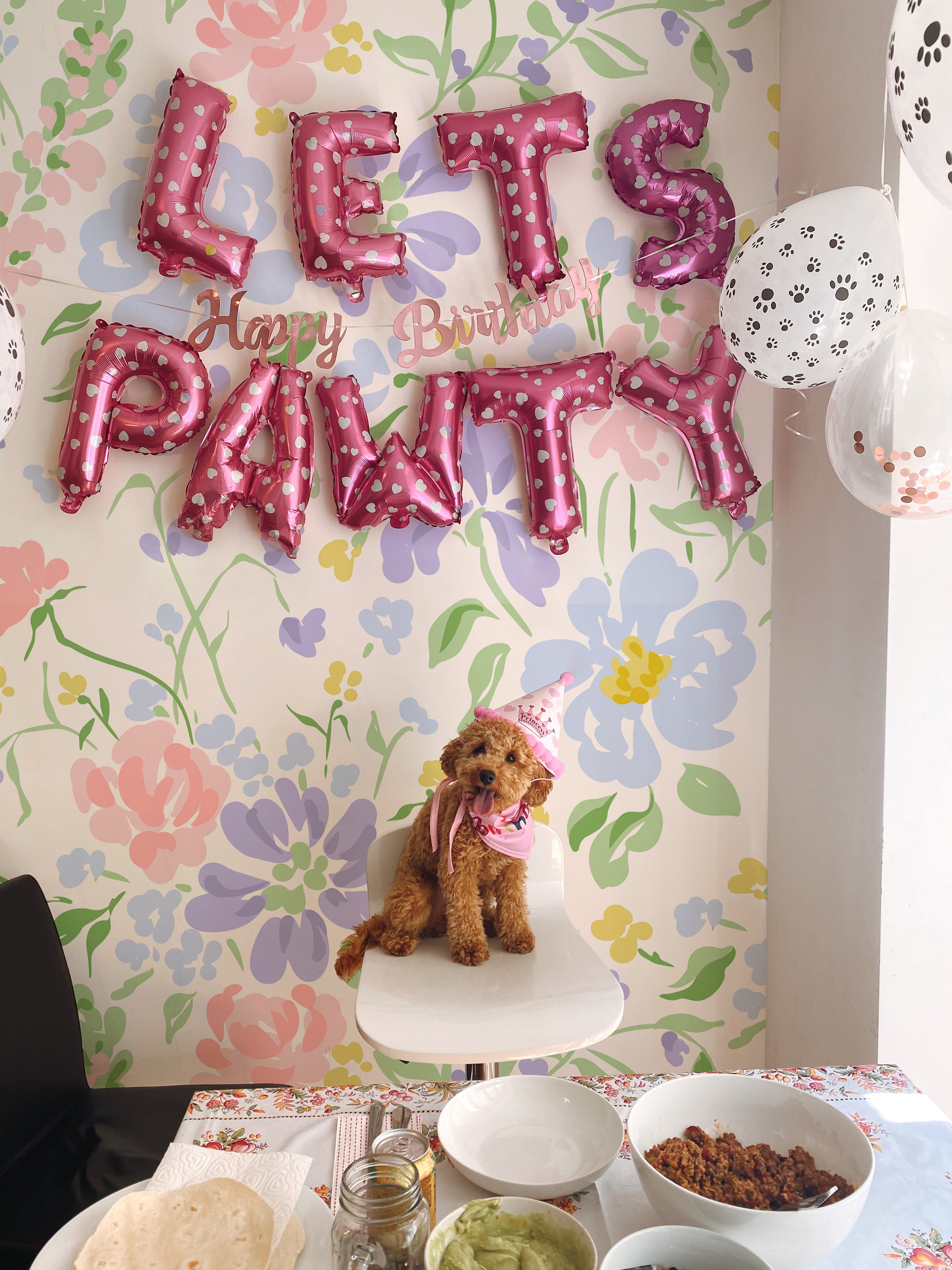 A festive birthday setting for a dog named 'Pawty' with colorful balloons spelling 'Let's Pawty Happy Birthday' against a backdrop of Romana Wallpaper. The wallpaper features a cheerful floral pattern in pastel shades of pink, blue, and yellow.