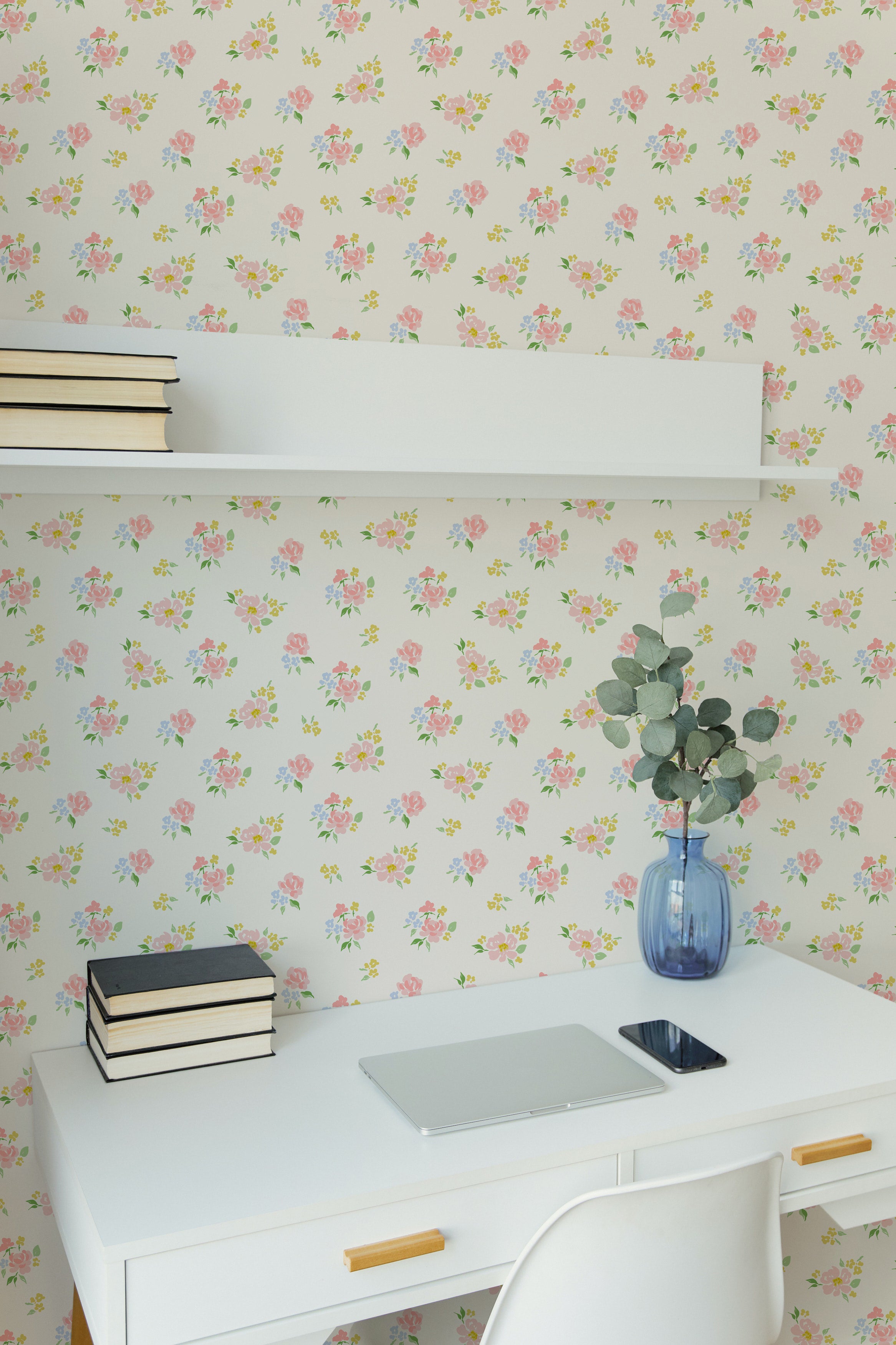 A modern workspace featuring the Rōma Hito Wallpaper, decorated with a delicate pattern of small pink and blue roses scattered across a light cream background. The scene includes a white desk with books and a blue vase with greenery, creating a fresh and inviting work environment.