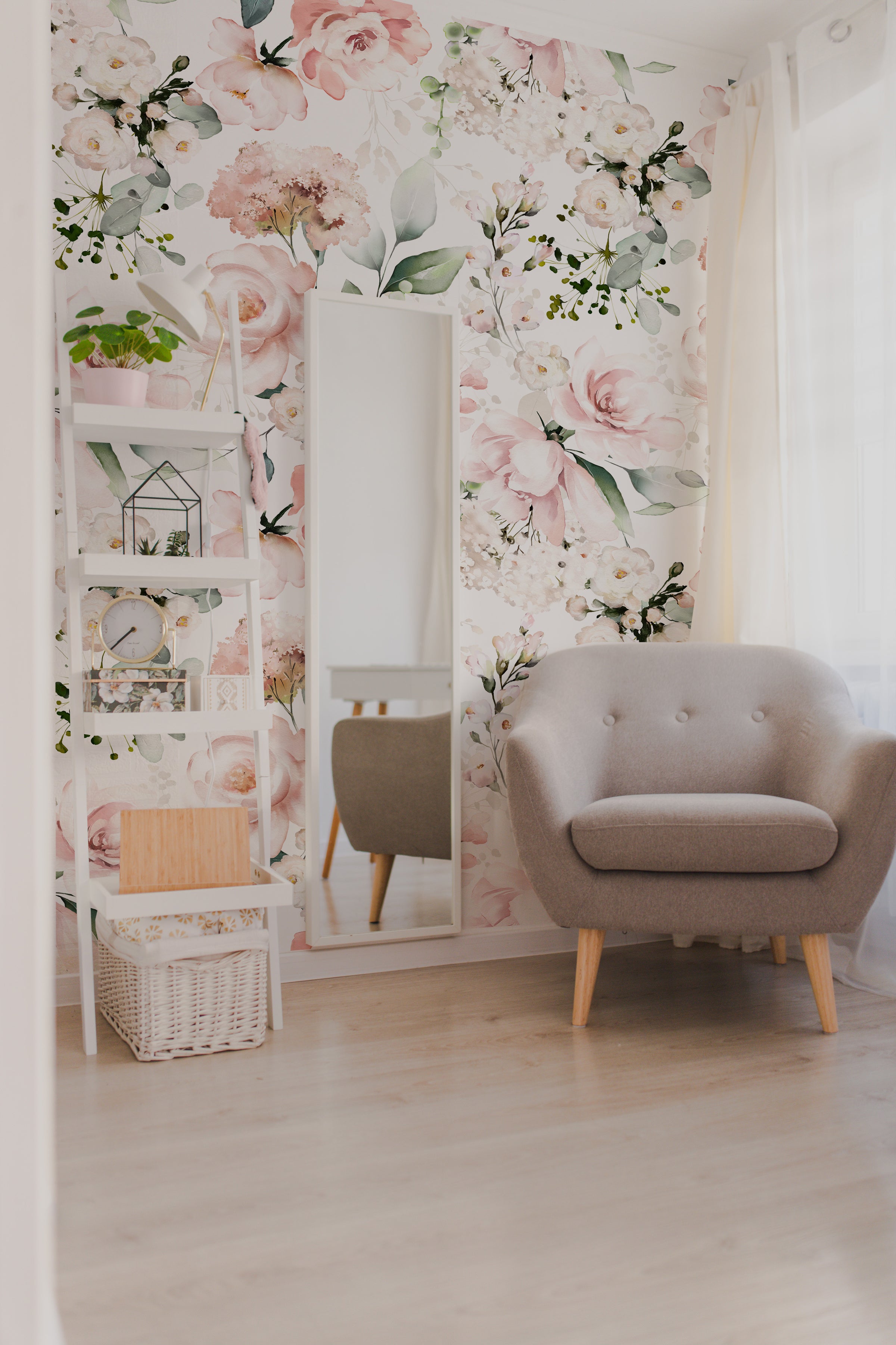 A cozy reading nook with the Pink Floral and Herbs Wallpaper - 50" creating a vibrant backdrop. The wall is adorned with large watercolor blooms in shades of pink and lush greenery. A white shelving unit decorated with plants and trinkets complements the wallpaper, beside a stylish grey armchair and a floor-length mirror that reflects the room's natural light.