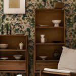 Cozy interior scene featuring Spring Pigeon Wallpaper with a rich botanical design of green leaves, white blossoms, and vibrant pigeons. A wooden bookshelf filled with ceramic pottery adds a rustic touch against the lively wallpaper background.