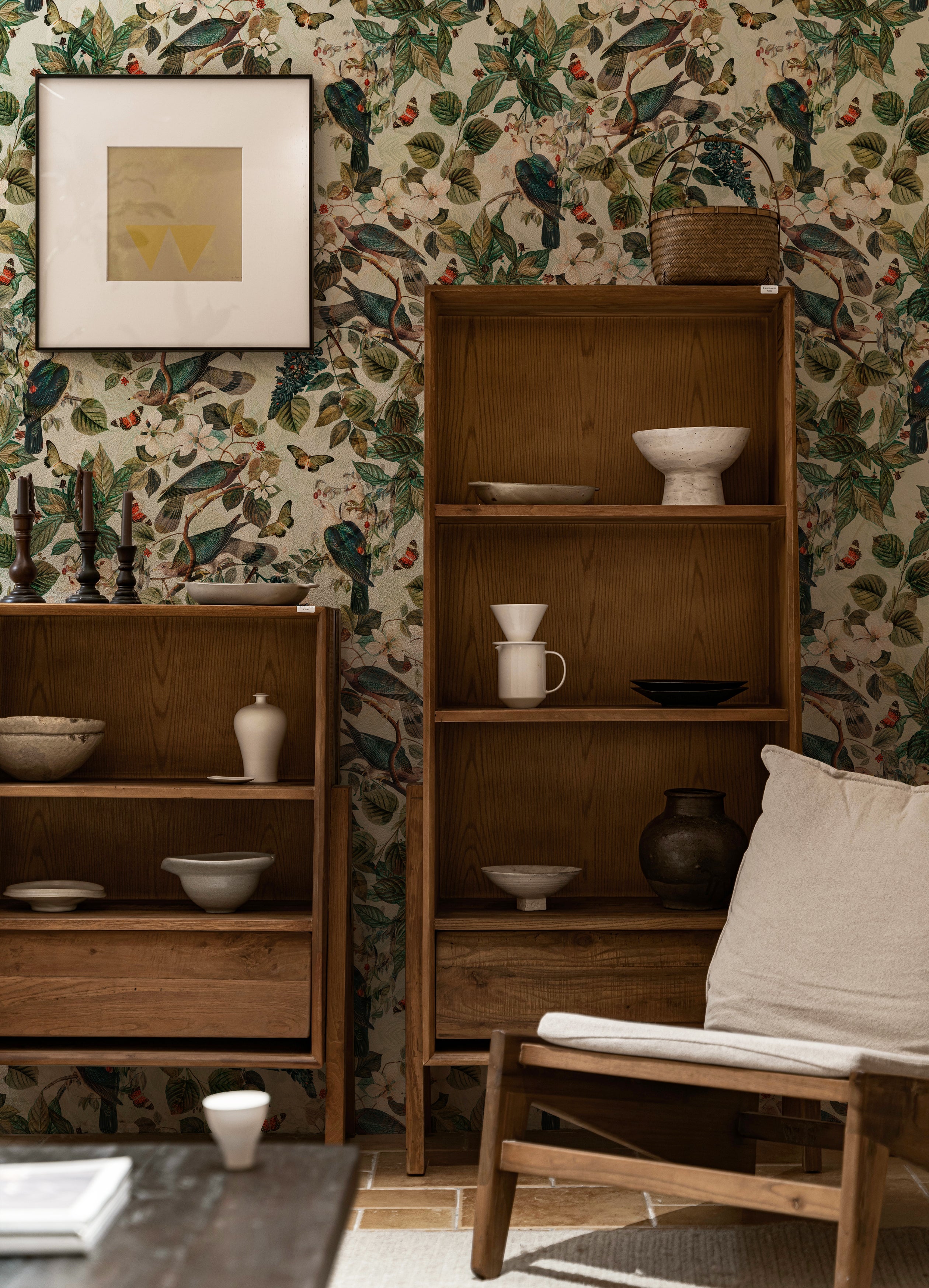Cozy interior scene featuring Spring Pigeon Wallpaper with a rich botanical design of green leaves, white blossoms, and vibrant pigeons. A wooden bookshelf filled with ceramic pottery adds a rustic touch against the lively wallpaper background.