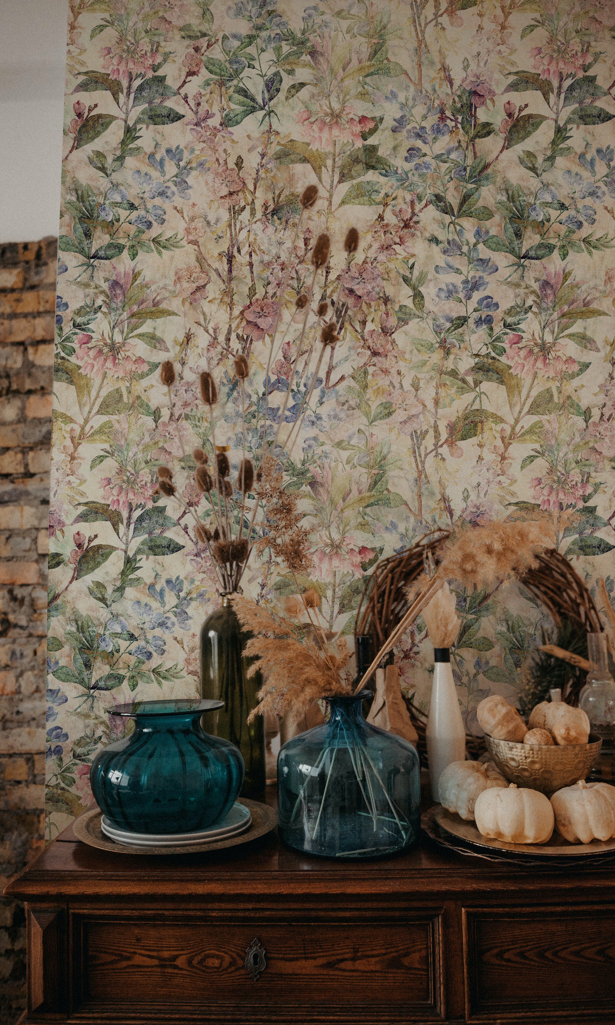 A close-up view of the "Ancient Florals Wallpaper" showcasing its intricate botanical print. The wallpaper decorates a room corner styled with dried flowers in vases and seasonal pumpkins, contributing to a rustic and quaint interior design.