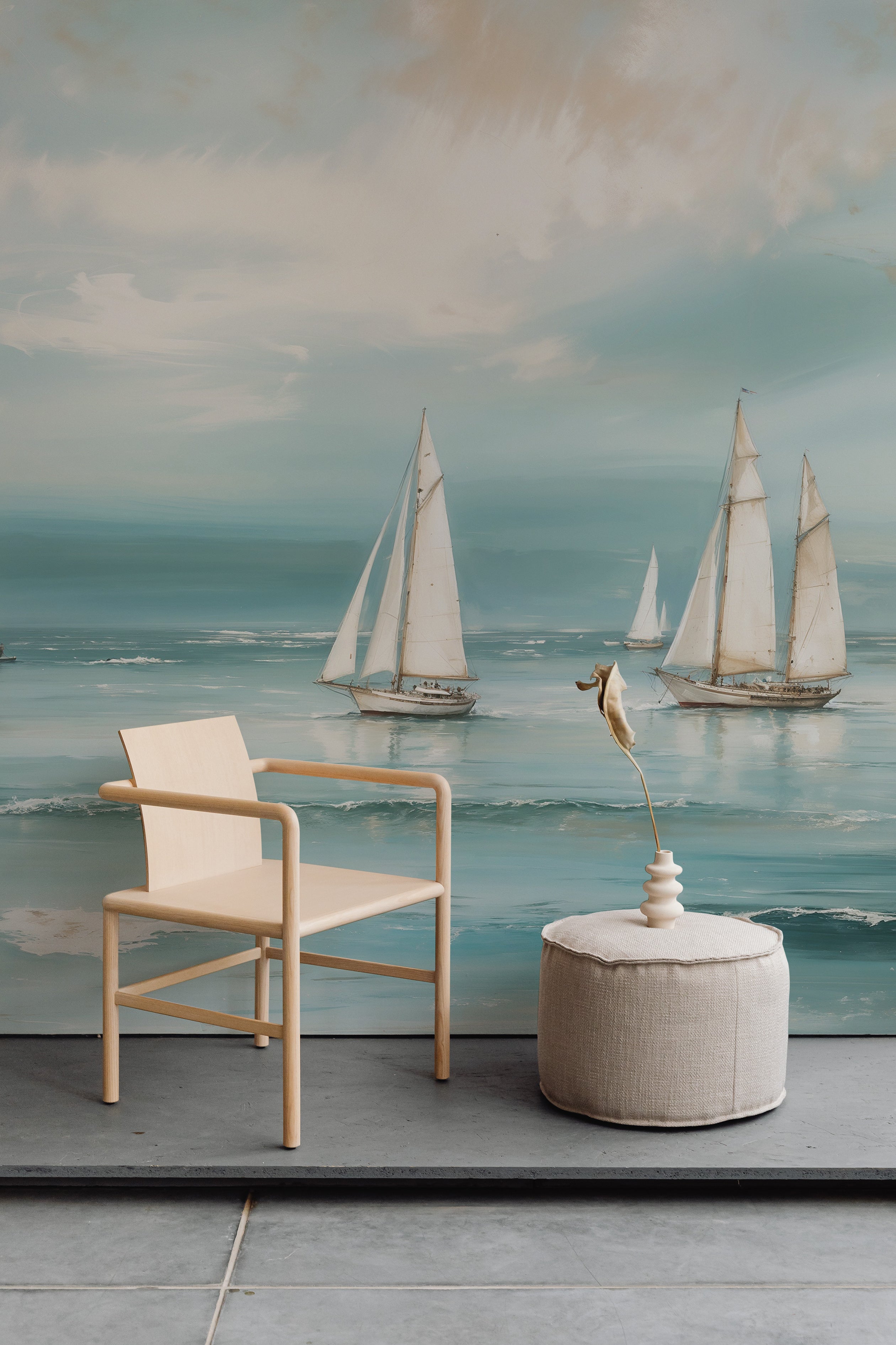 A serene and stylish room with a Seaside Wallpaper Mural depicting a beautiful ocean scene with sailboats. The mural creates a tranquil backdrop to a minimalistic seating area featuring a light wooden chair, a circular ottoman, and a delicate sculptural ornament.
