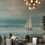A cafe setting with the Seaside Wallpaper Mural providing a calming ocean view with sailboats. The area is furnished with plush gray chairs and wooden tables, under a magnificent chandelier, creating an elegant and peaceful dining experience.