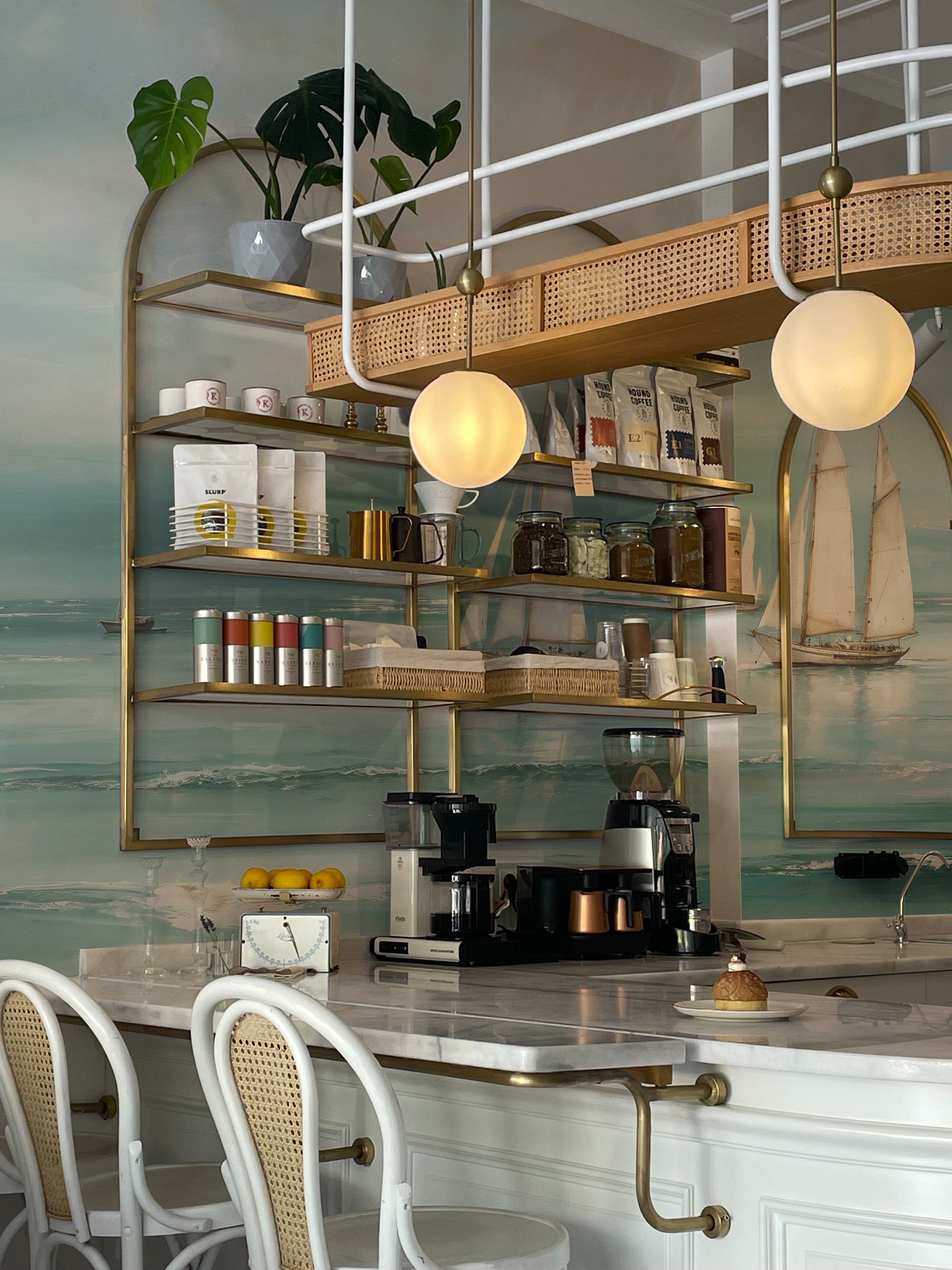 An inviting coffee bar against the Seaside Wallpaper Mural showcasing sailboats on a vivid sea. The bar is equipped with modern appliances and open shelving filled with coffee supplies and plants, enhancing the fresh, nautical theme.