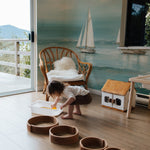 A child's play area enhanced by the Seaside Wallpaper Mural, featuring sailboats and a dreamy seascape. A young child plays on the floor, adding a lively and personal touch to the serene oceanic backdrop.