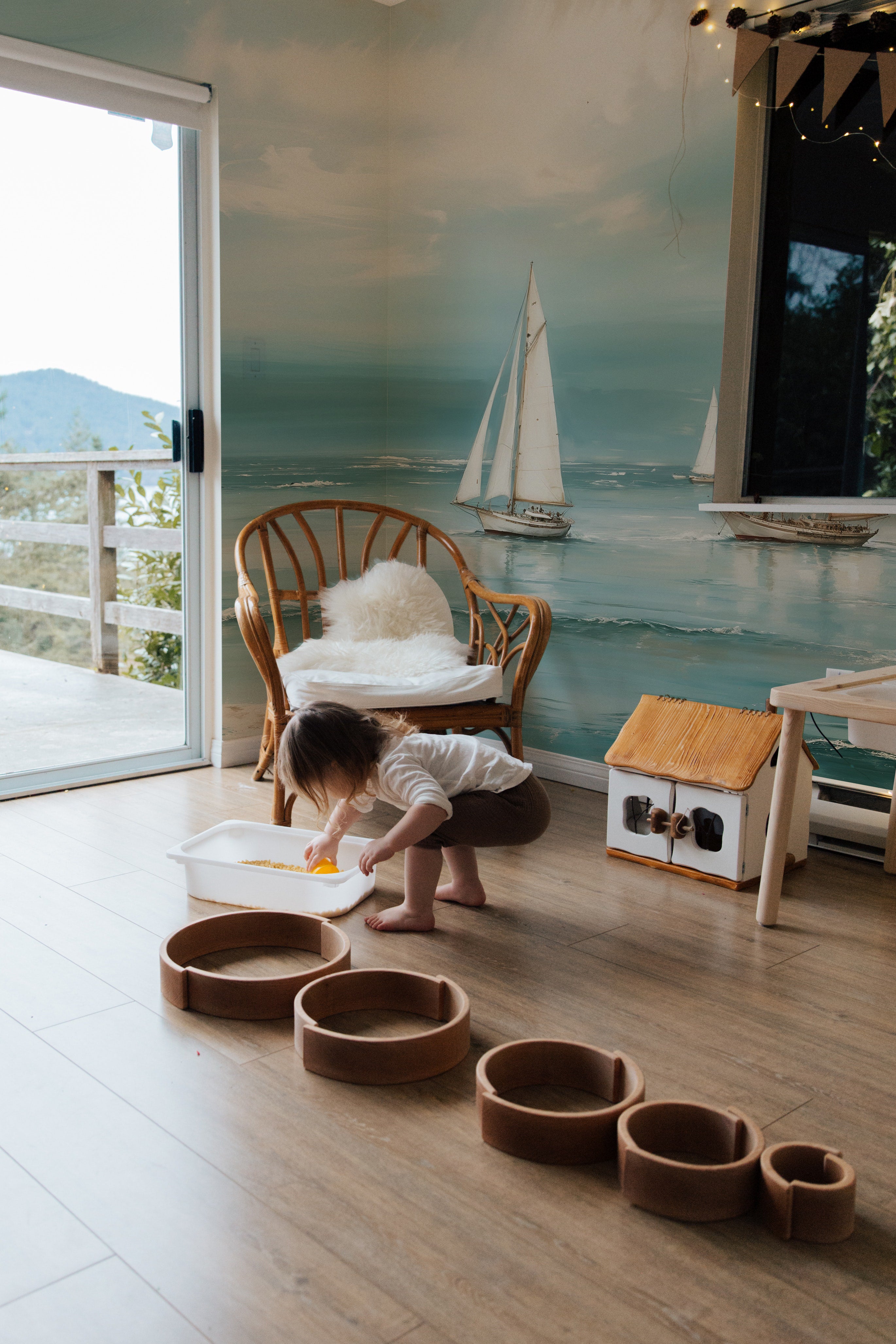 A child's play area enhanced by the Seaside Wallpaper Mural, featuring sailboats and a dreamy seascape. A young child plays on the floor, adding a lively and personal touch to the serene oceanic backdrop.