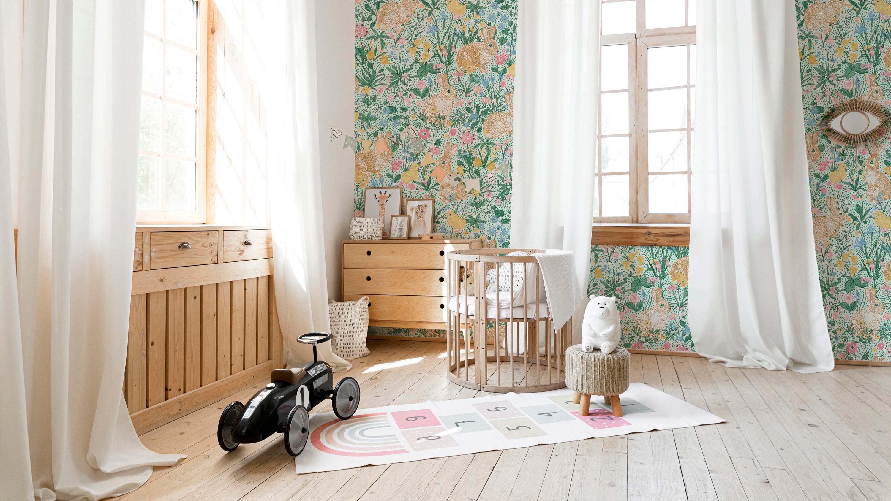 "Bright and airy children's room featuring a colorful floral wallpaper with rabbits, birds, and a variety of flowers. The room includes wooden furniture, a circular crib, and playful decor items like a small ride-on car and a hopscotch rug.
