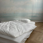 A serene bedroom featuring a wall mural depicting a calm seascape with multiple sailing ships in the distance under a cloud-filled sky. The room has a minimalist design with a white bedspread and hardwood floor.