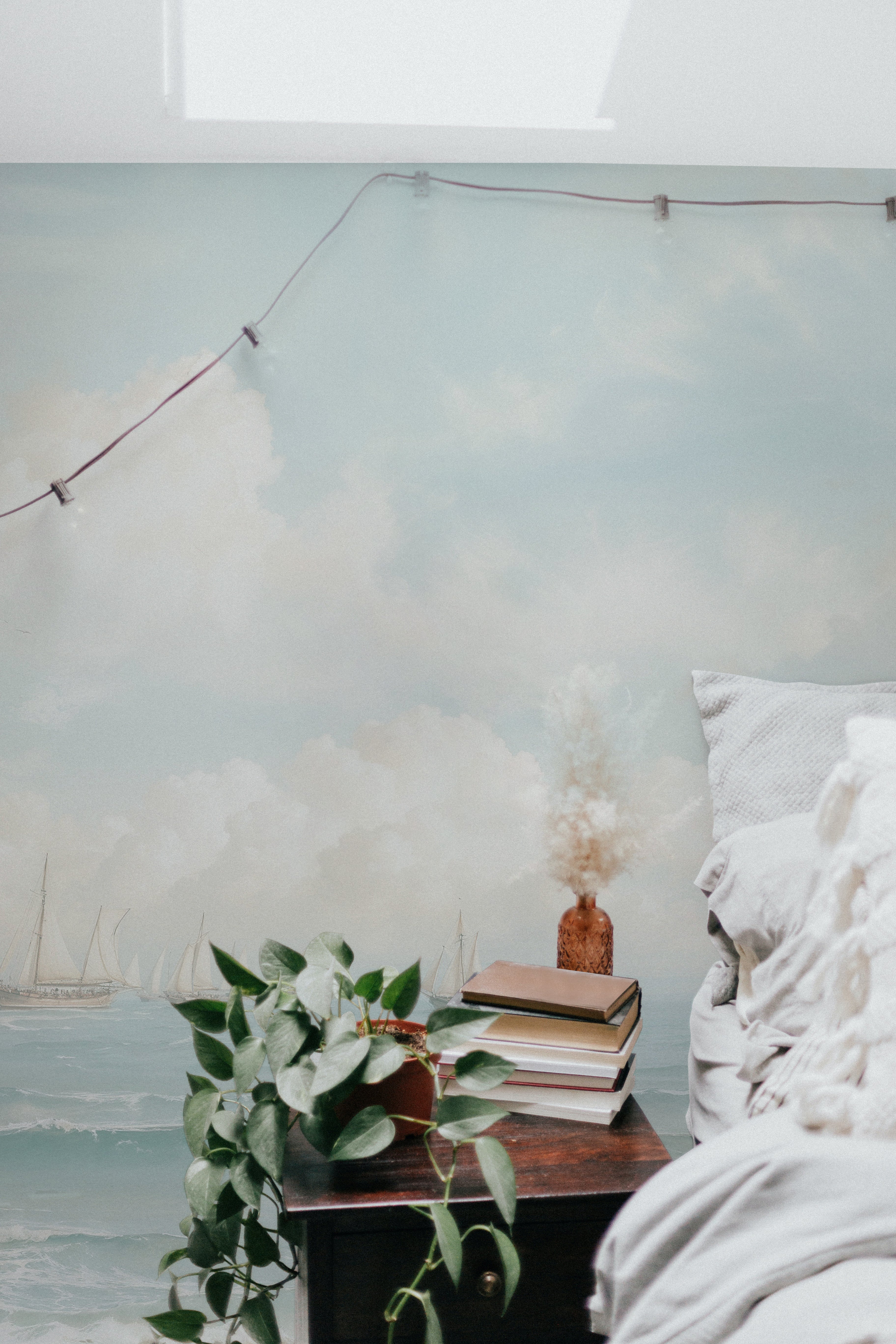 A cozy corner of a room with a detailed seascape wall mural displaying sailboats. The setting includes a side table with books and a plant, next to a bed adorned with light-colored linens.