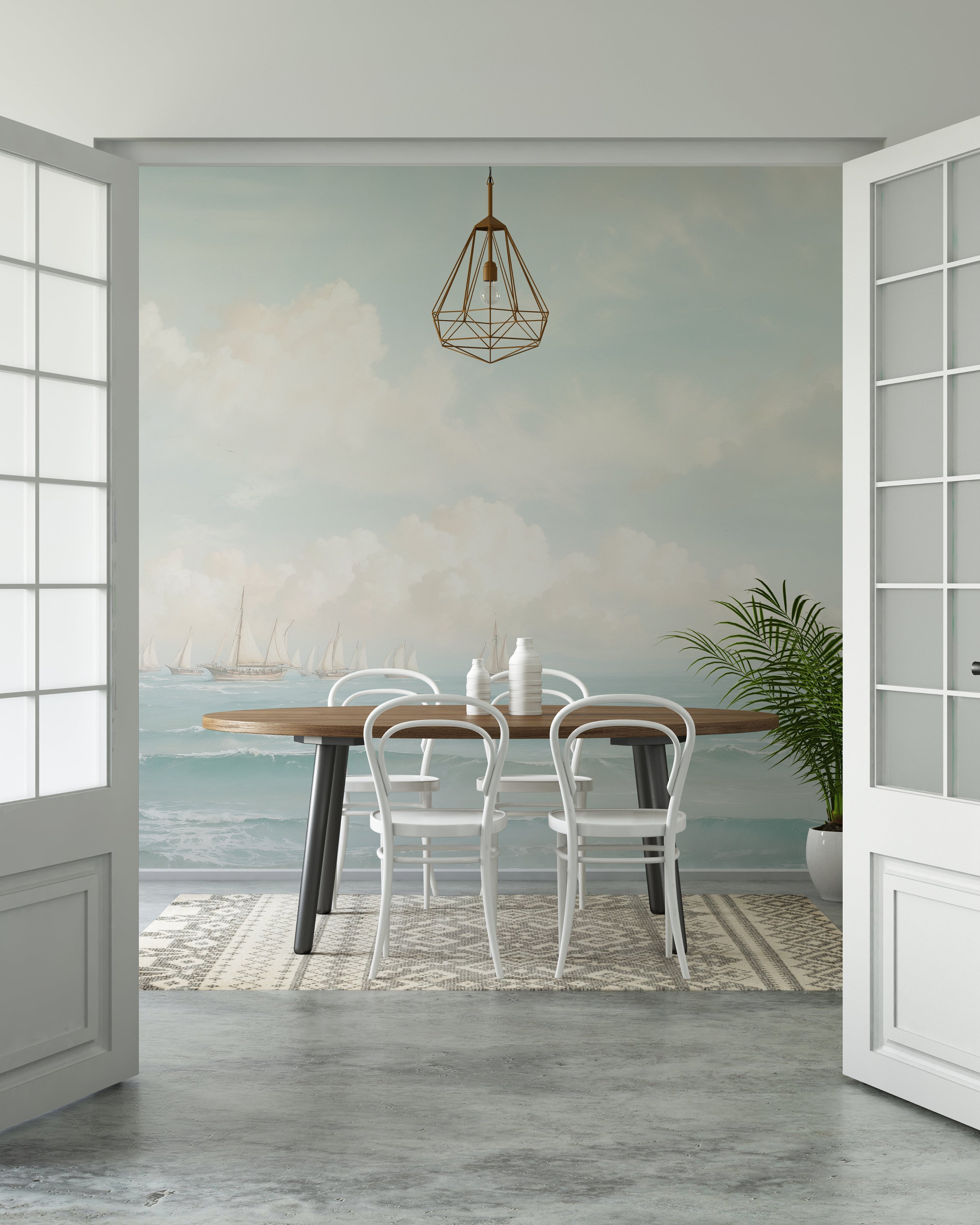 A dining area with modern furnishings set against a large wall mural of a light and airy seascape featuring distant sailboats. The scene includes a hanging geometric light fixture and a pair of glass doors filling the room with natural light.