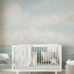 A nursery with walls adorned by a seascape mural showing sailing ships on a gentle sea. The room is decorated in white and grey tones, with a modern crib, soft textures, and playful elements like alphabet blocks on the floor.