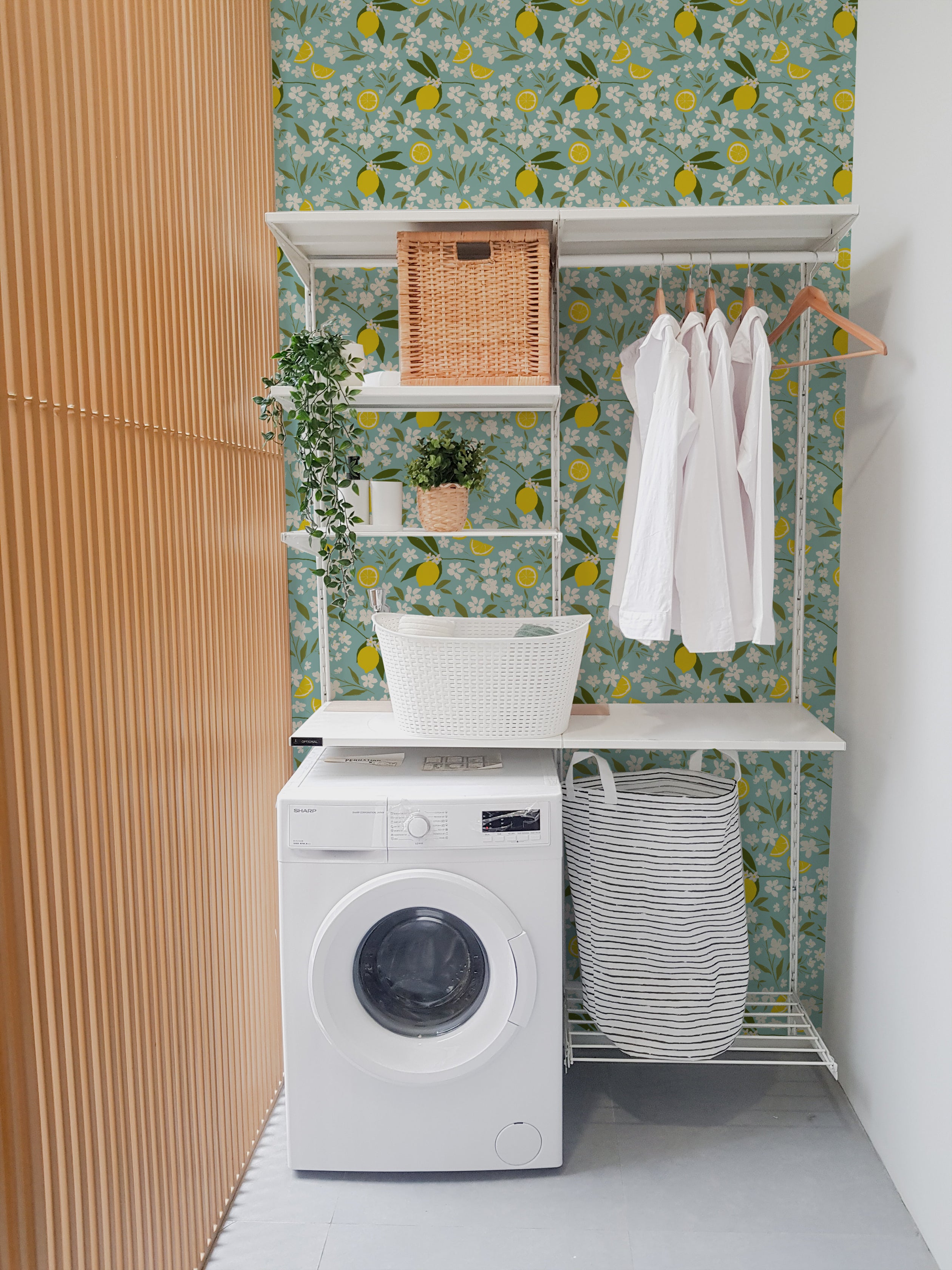 A laundry area enhanced by the Lemon + Jasmine Wallpaper, showcasing a lively pattern of yellow lemons and white jasmine flowers on a light blue background. The room includes white shelving with various laundry essentials and a washing machine, exuding a clean and cheerful vibe.