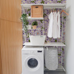 A beautifully decorated laundry room featuring the Passion Flower Wallpaper with a lavender background adorned with vibrant floral patterns of blue, white, and yellow flowers. The room is complemented by a modern washing machine, wooden shelving filled with linens and decorative items, and woven baskets, creating a charming and functional space.