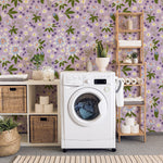 A beautifully decorated laundry room featuring the Passion Flower Wallpaper with a lavender background adorned with vibrant floral patterns of blue, white, and yellow flowers. The room is complemented by a modern washing machine, wooden shelving filled with linens and decorative items, and woven baskets, creating a charming and functional space.