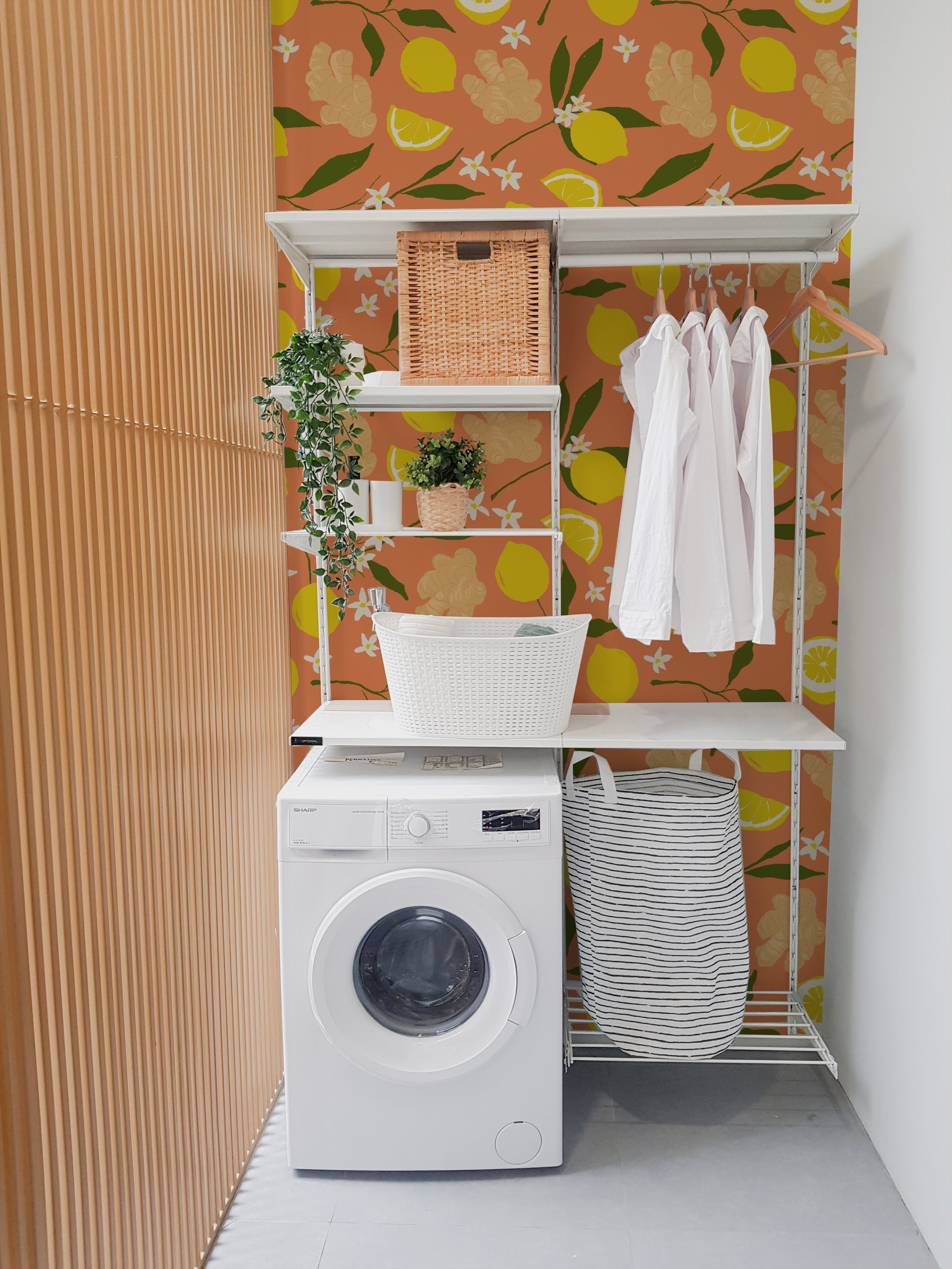 A cozy corner of a laundry room enhanced by the Lemon Ginger Wallpaper. This bright pattern consists of yellow lemons, ginger flowers, and small white blossoms on an orange background. The room features a washing machine, white shelving with wicker baskets, and neatly hung white bathrobes, adding a touch of warmth and organization.