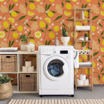 A vibrant laundry room featuring the Lemon Ginger Wallpaper, adorned with a pattern of yellow lemons, ginger flowers, and small white blossoms set against a rich orange backdrop. The scene includes a white washing machine, woven baskets, and wooden shelves filled with laundry essentials and decorative plants, creating a cheerful and functional space.