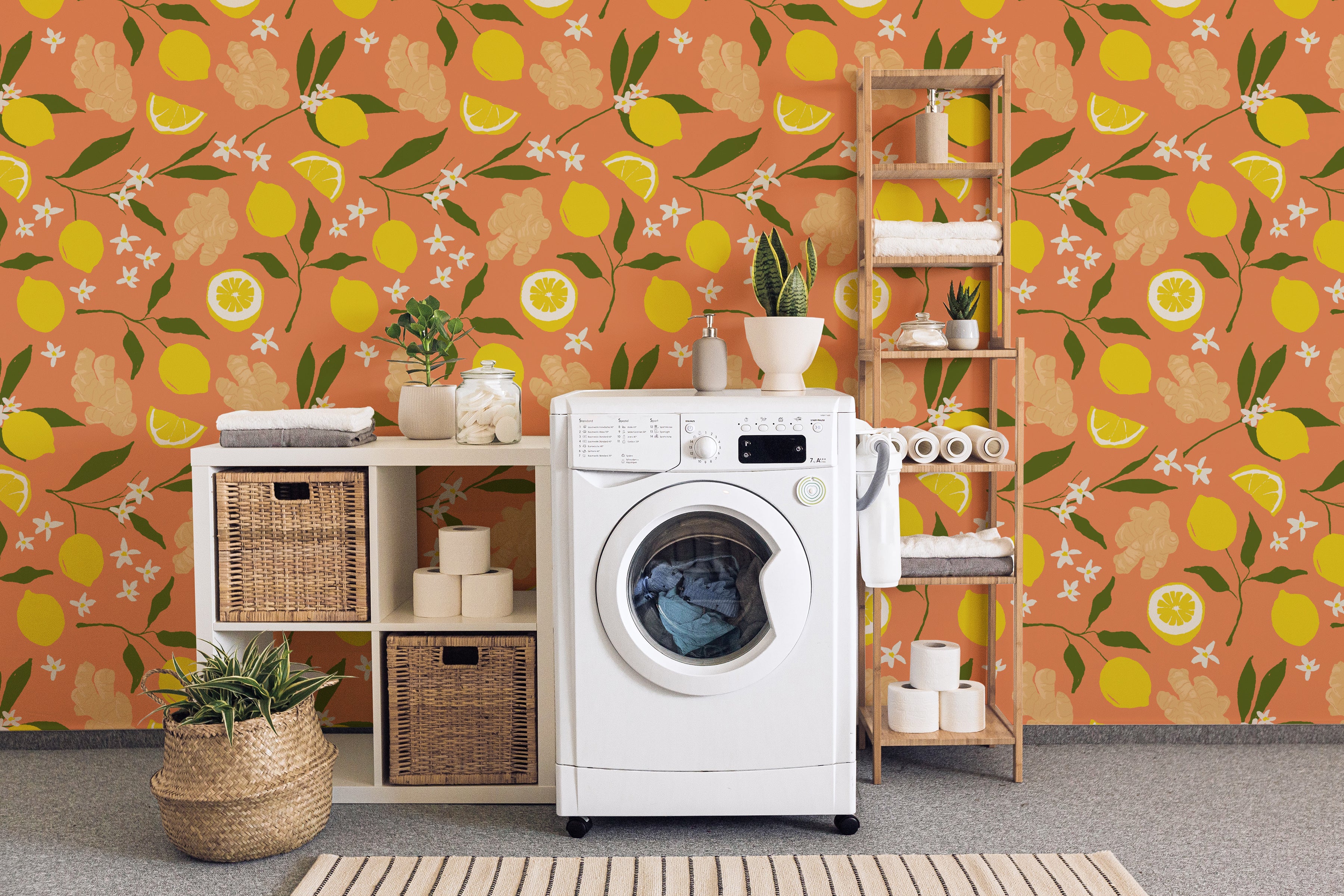 A vibrant laundry room featuring the Lemon Ginger Wallpaper, adorned with a pattern of yellow lemons, ginger flowers, and small white blossoms set against a rich orange backdrop. The scene includes a white washing machine, woven baskets, and wooden shelves filled with laundry essentials and decorative plants, creating a cheerful and functional space.