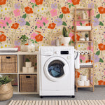 Colorful floral wallpaper in a small laundry room with a washing machine, shelves holding laundry baskets, and hanging white robes. The wallpaper has a vibrant pattern of large red, pink, and purple flowers mixed with white daisies against a beige background."