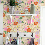 Brightly colored floral wallpaper in a bathroom setting with a white shelf holding various toiletries and a plant. The wall features large orange, pink, and violet flowers with smaller daisies and decorative dots on a beige background.