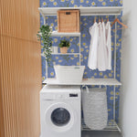 "Bright and cheerful laundry room with light blue walls covered in a floral wallpaper featuring white and yellow daisies, a white washing machine, white shelving units with various household items, and wicker baskets.