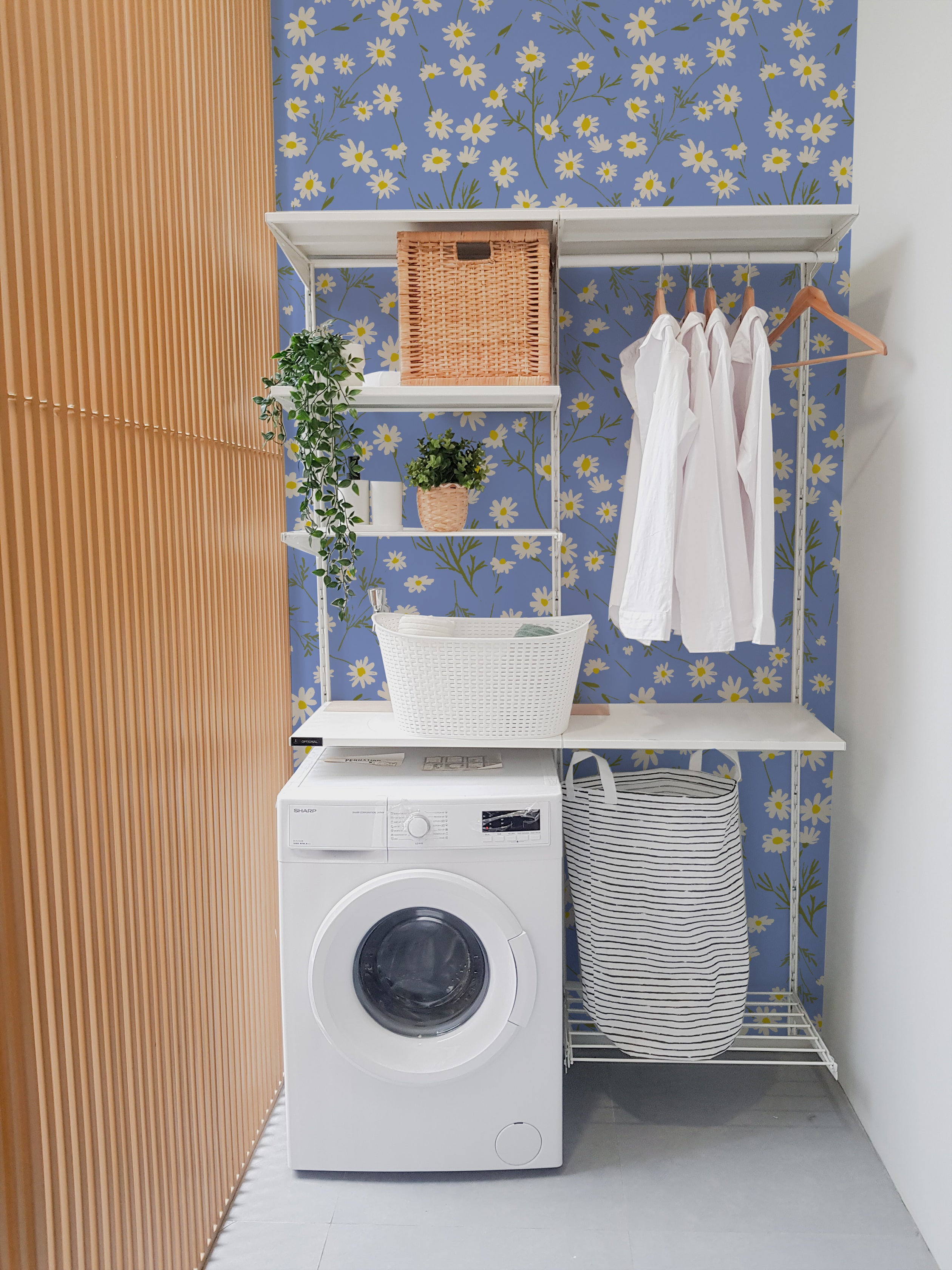 "Bright and cheerful laundry room with light blue walls covered in a floral wallpaper featuring white and yellow daisies, a white washing machine, white shelving units with various household items, and wicker baskets.