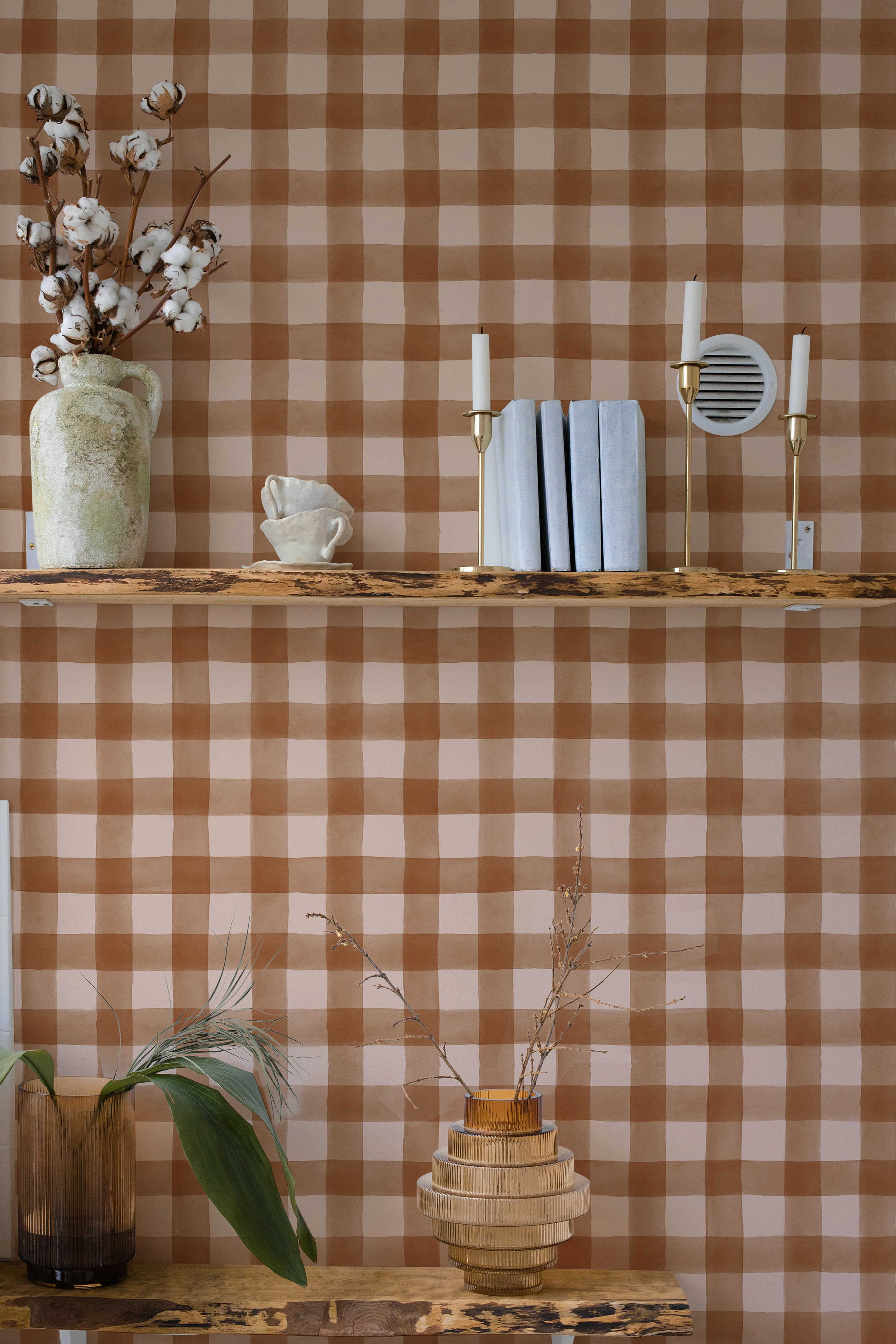 A warmly styled interior scene featuring a wall covered in Cuadro de Búfalo wallpaper with a classic brown and beige checkered pattern. The decor includes a rustic wooden shelf displaying a vase with cotton branches, a cup shaped like a spilt drink, and modern white candle holders.