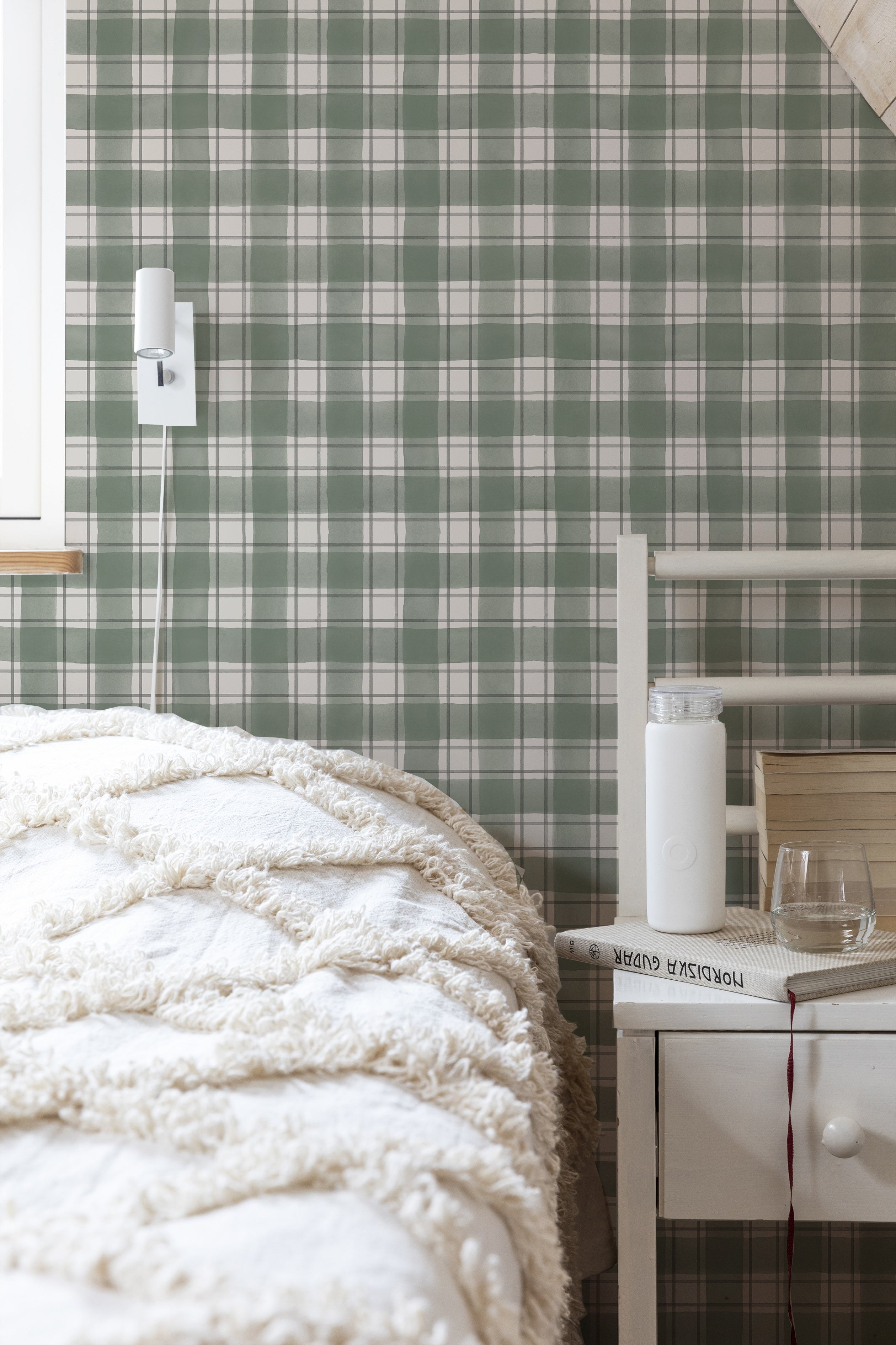 A cozy bedroom featuring a green buffalo check pattern on the walls. The room is furnished with a fluffy white comforter on the bed, a minimalist white nightstand, and a small lamp.