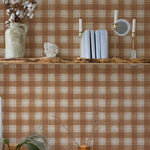An elegantly styled home interior with a rust-colored buffalo check pattern wall. Decor includes a ceramic vase with cotton branches, candle holders, and neatly stacked books on a wooden shelf.