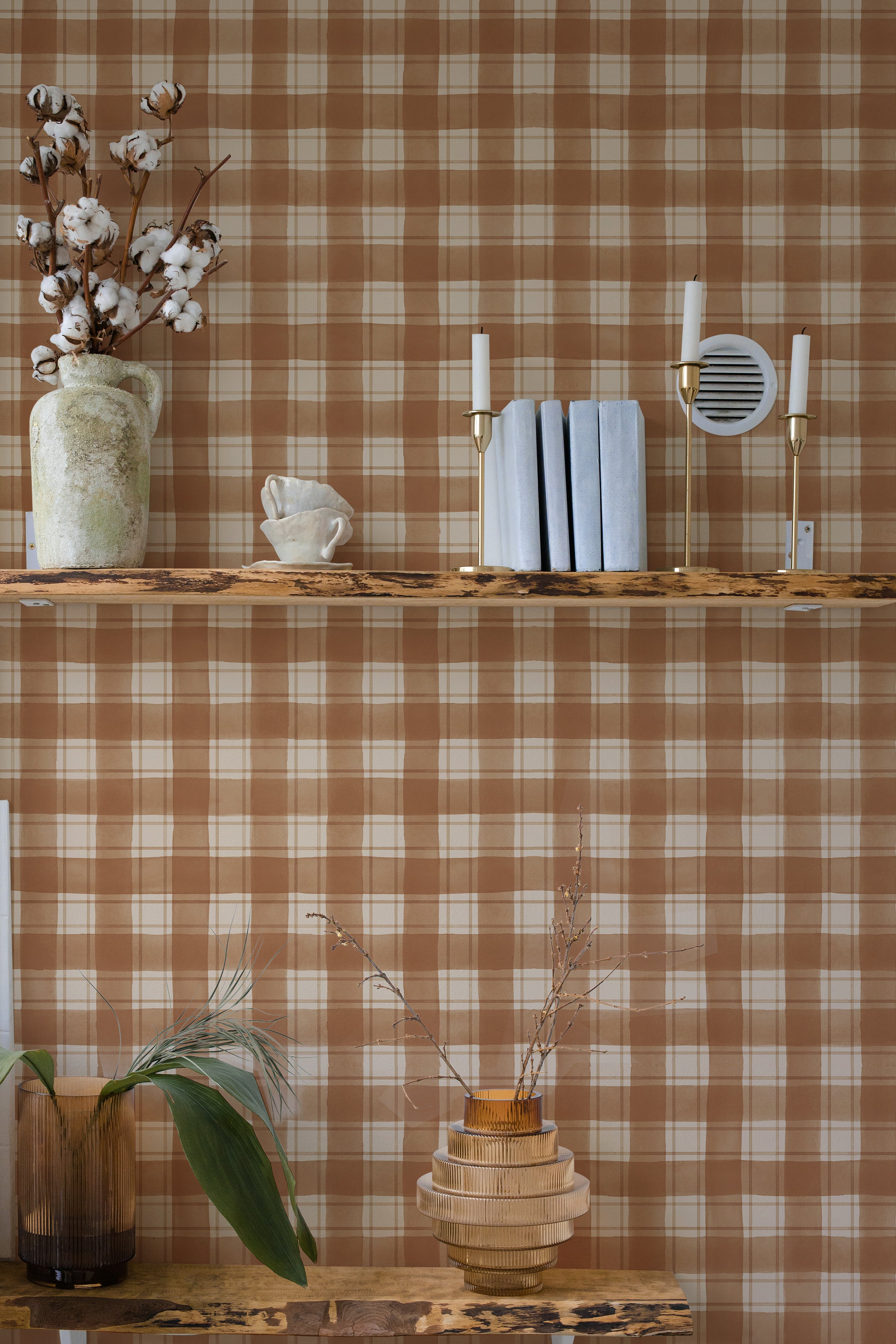 An elegantly styled home interior with a rust-colored buffalo check pattern wall. Decor includes a ceramic vase with cotton branches, candle holders, and neatly stacked books on a wooden shelf.